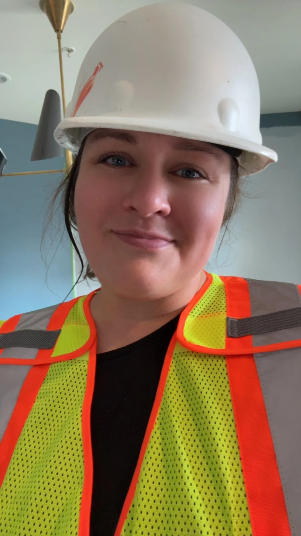 A woman wearing a yellow reflective safety vest and a white construction helmet, smiling indoors.
