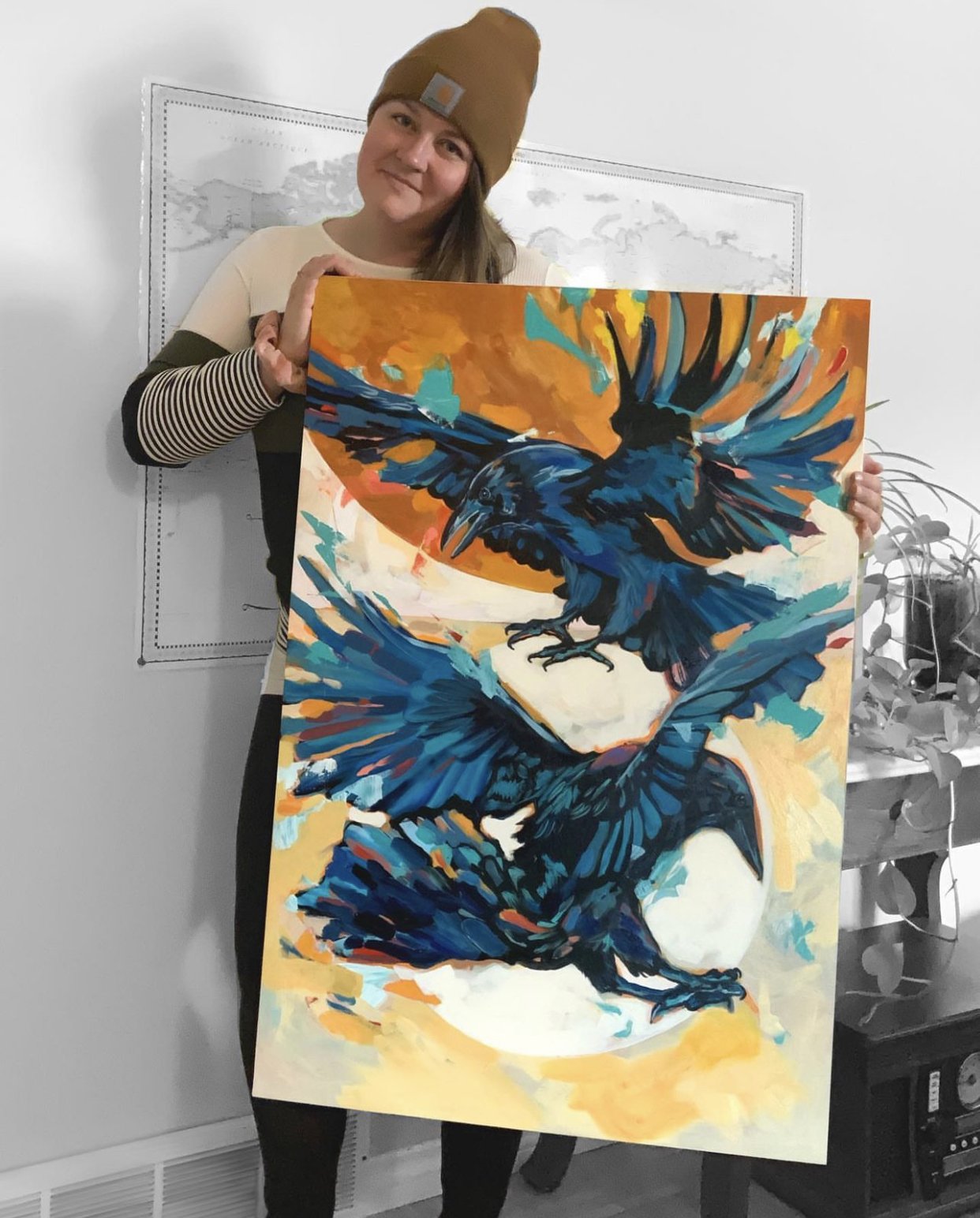 A woman wearing a brown beanie and a striped shirt holding a colorful painting of three black ravens with spread wings against an orange and yellow background.