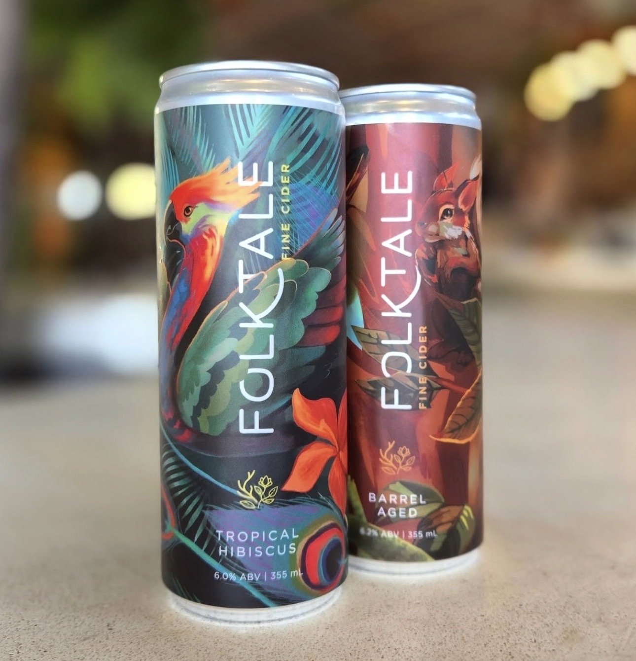 Two cans of Folktale fine cider on a blurred outdoor background. One can features a colorful tropical hibiscus design with a Fenghuang bird parrot, and the other has a barrel aged theme with a squirrel figure in apple trees.