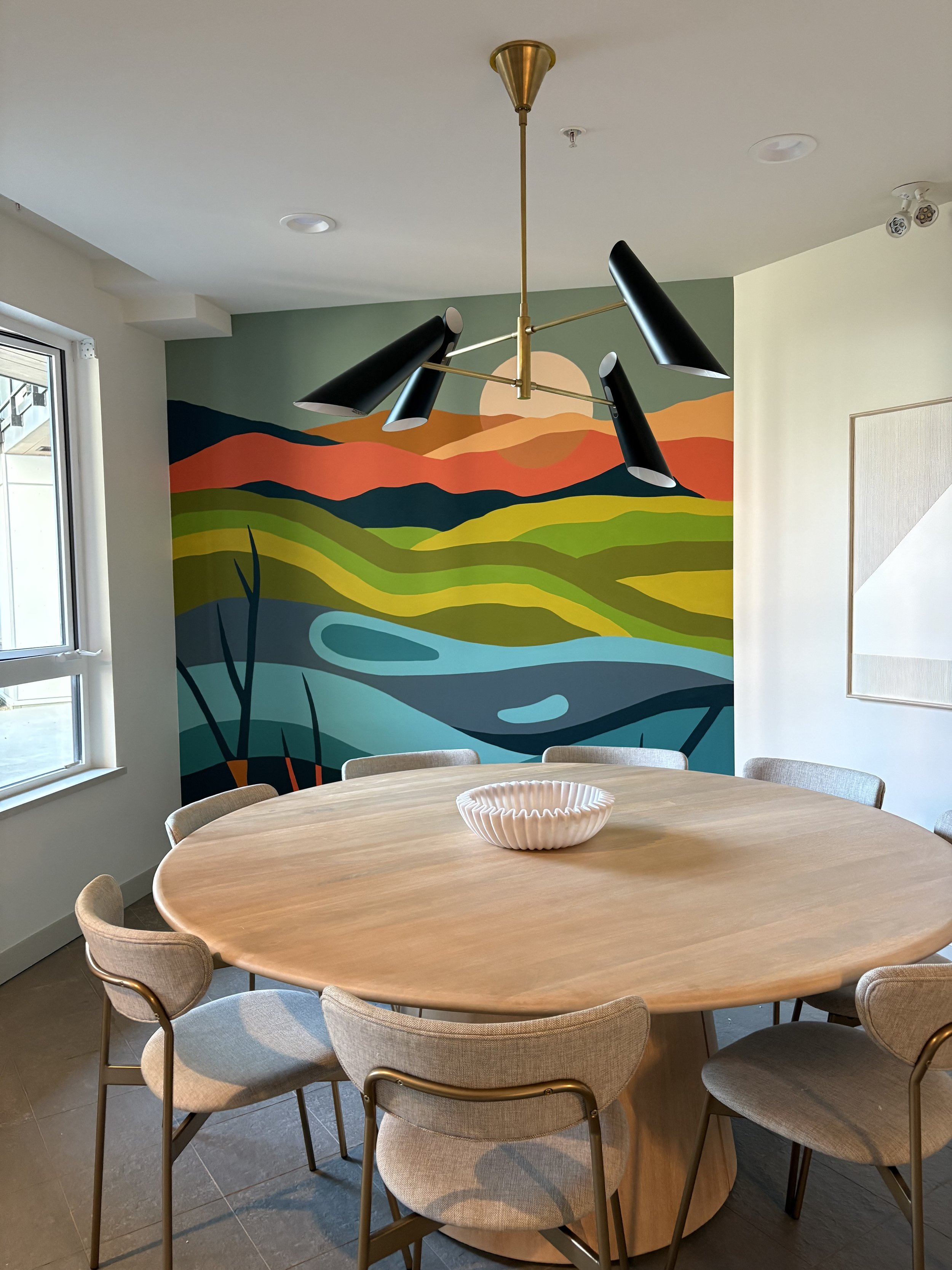 A round wooden table surrounded by beige chairs, with a decorative bowl in the center, inside a room with a colorful mural of a landscape on the wall and a modern black and gold chandelier overhead.