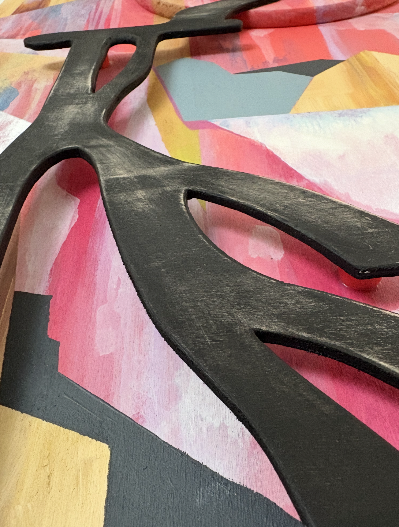 Close-up photo of black decorative wall art with geometric design, placed on a colorful abstract painted surface with pink, yellow, blue, and white hues.