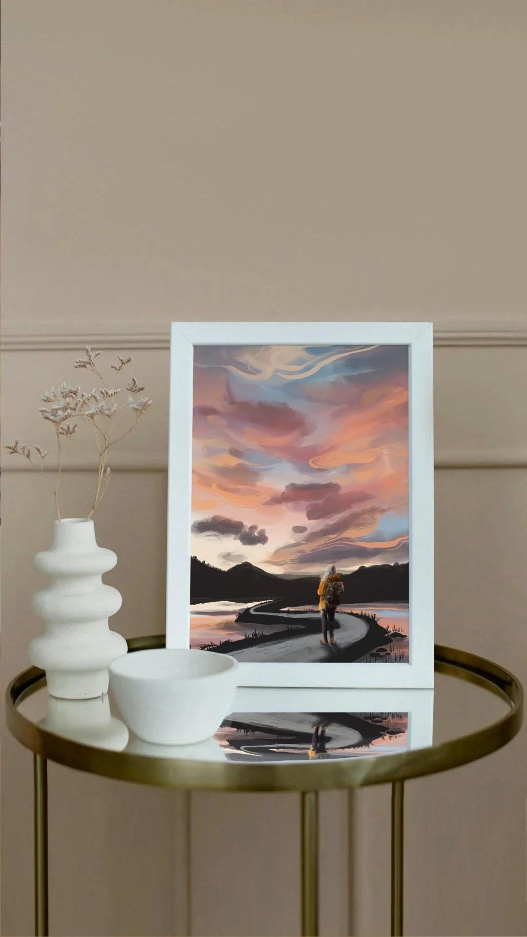 A framed landscape painting of a person walking along a winding road at sunset, with colorful sky and dark mountains in the background, placed on a round gold table next to a white vase with dried flowers and a white ceramic bowl.