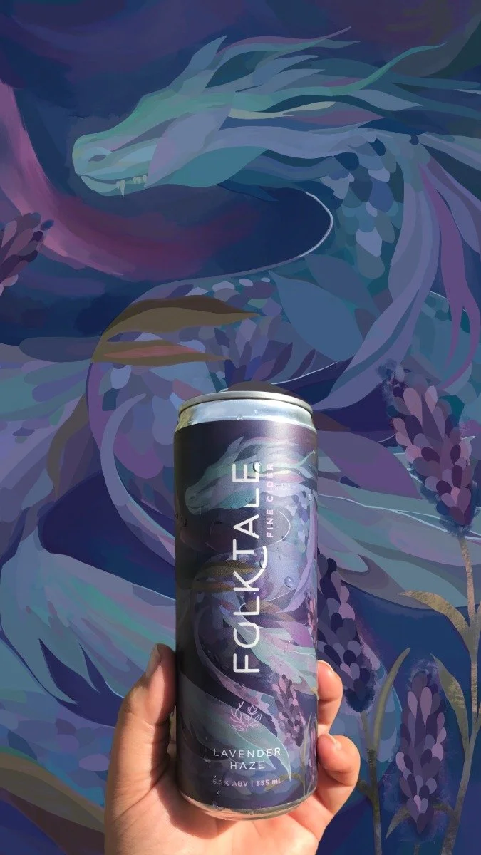 A hand holding a can of Folktale Lavender Haze sparkling water in front of a watercolor mural with a dragon and floral elements in shades of blue, purple, and teal.