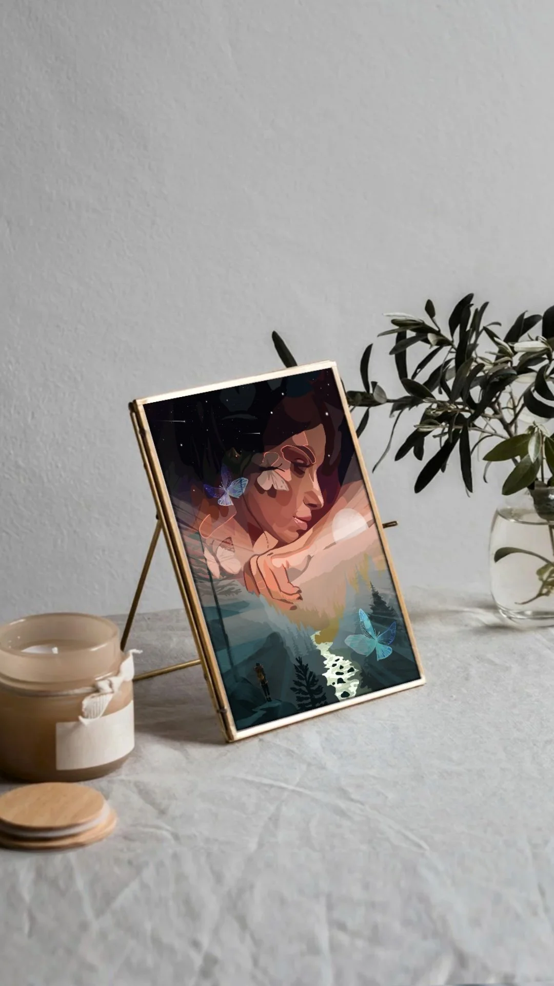 Art print of a woman with closed eyes, surrounded by butterflies and natural elements, displayed on a gold stand on a white textured surface with a small plant in a glass vase and candles nearby.