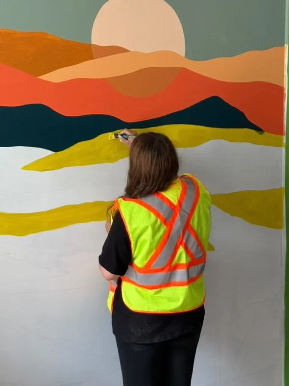 A woman in a neon yellow safety vest painting a mural of a sunset landscape with mountains and water.
