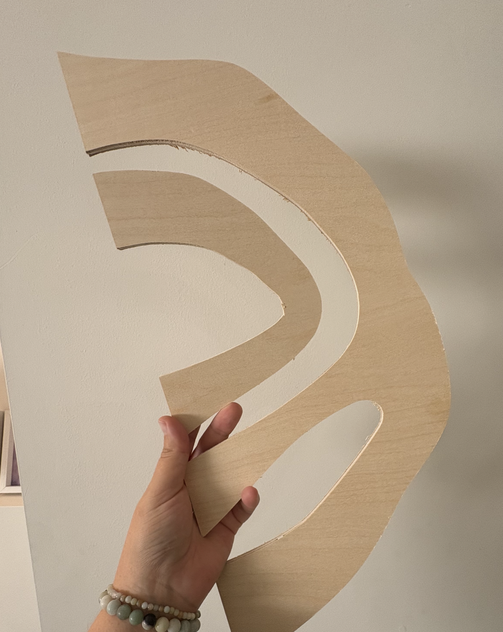 A person holding a large wooden cutout against a beige wall.