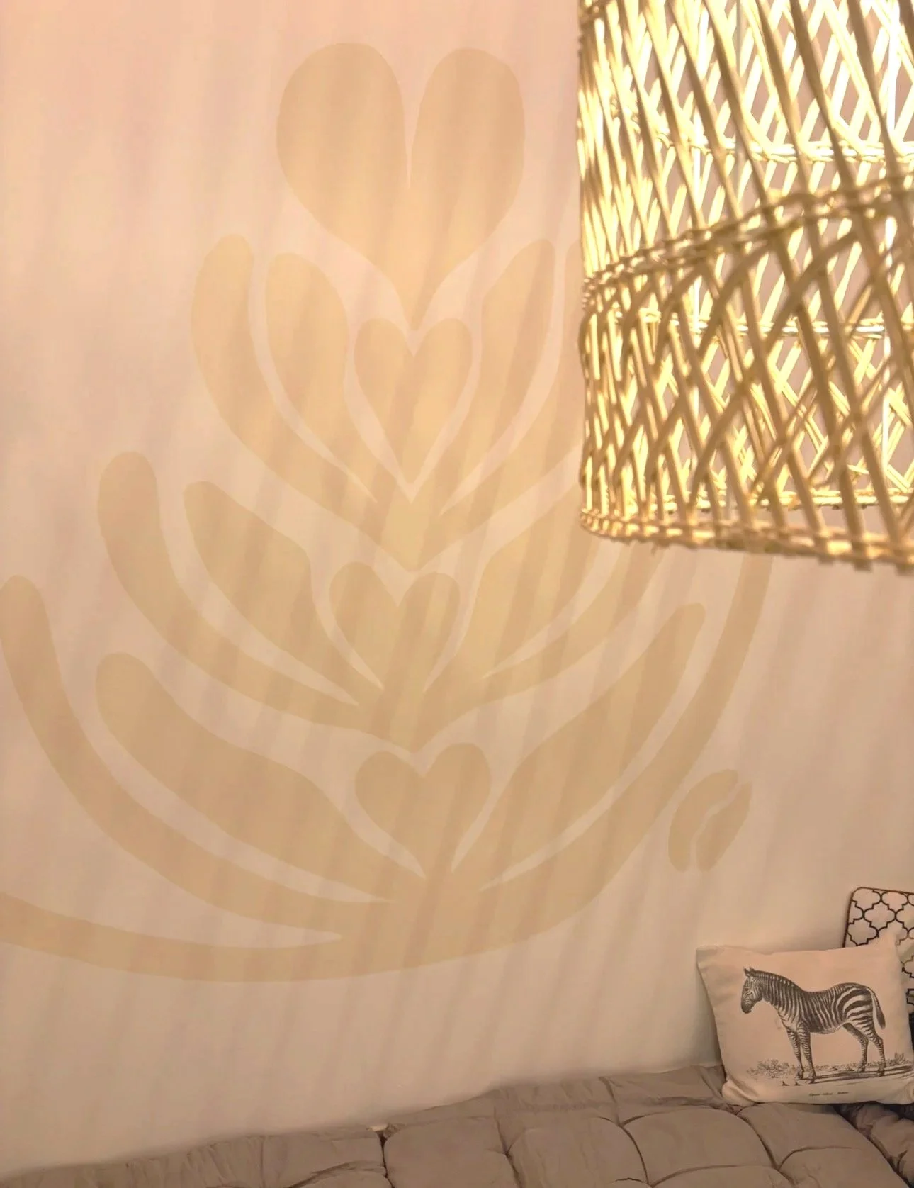 A beige wall with a large, stylized floral pattern in a slightly darker beige. To the right, there is a woven lampshade with warm lighting. At the bottom, part of a beige tufted sofa and a pillow with a zebra print are visible. Bright Jenny Coffee,