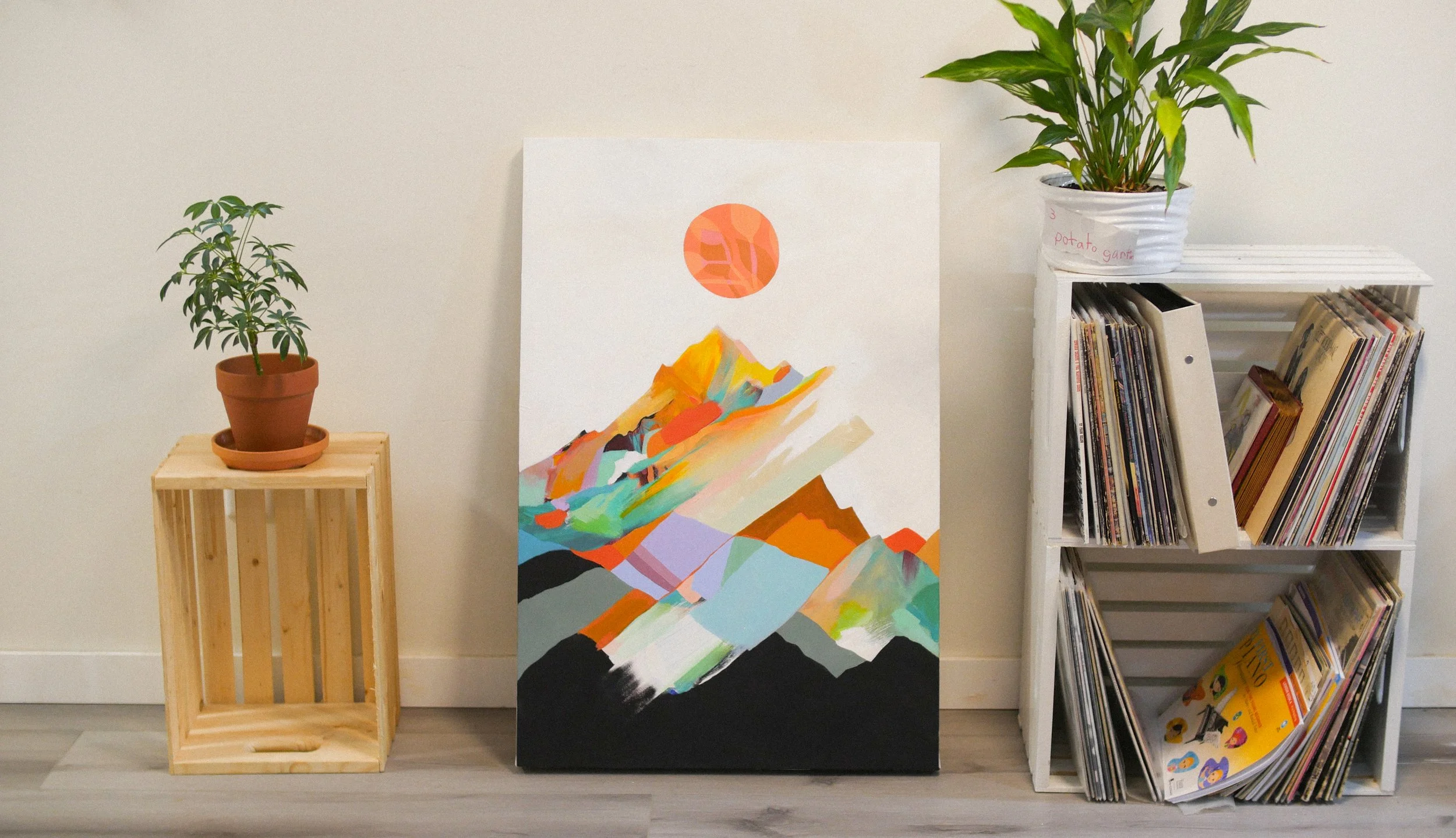 Decorative interior with a wooden crate holding a potted plant on the left, a colorful abstract mountain painting in the center, and a white bookshelf with vinyl records and a potted plant on top on the right.