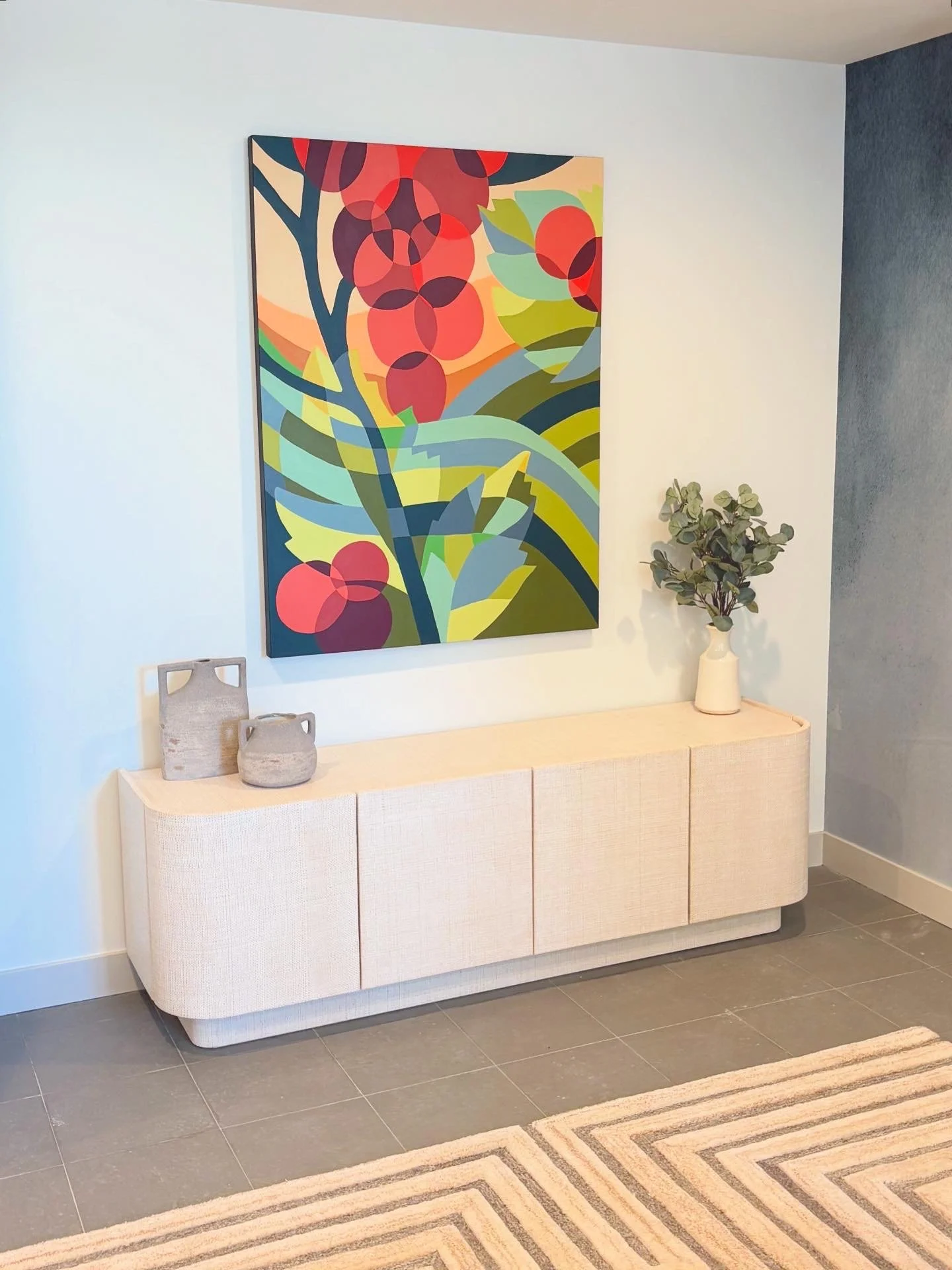 A beige sideboard with rounded edges and textured surface, decorated with two gray ceramic vases and a tall white vase with green leafy plant, against a light wall with a colorful abstract painting of botanical shapes hanging above.