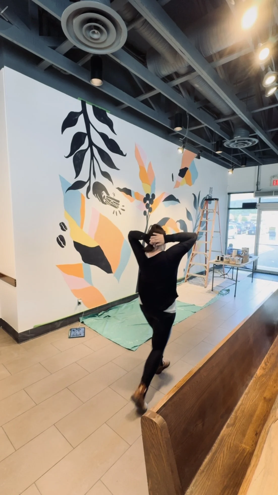 Interior of a cafe or store with a large abstract wall mural and a person walking past, with a ladder and table with supplies nearby.