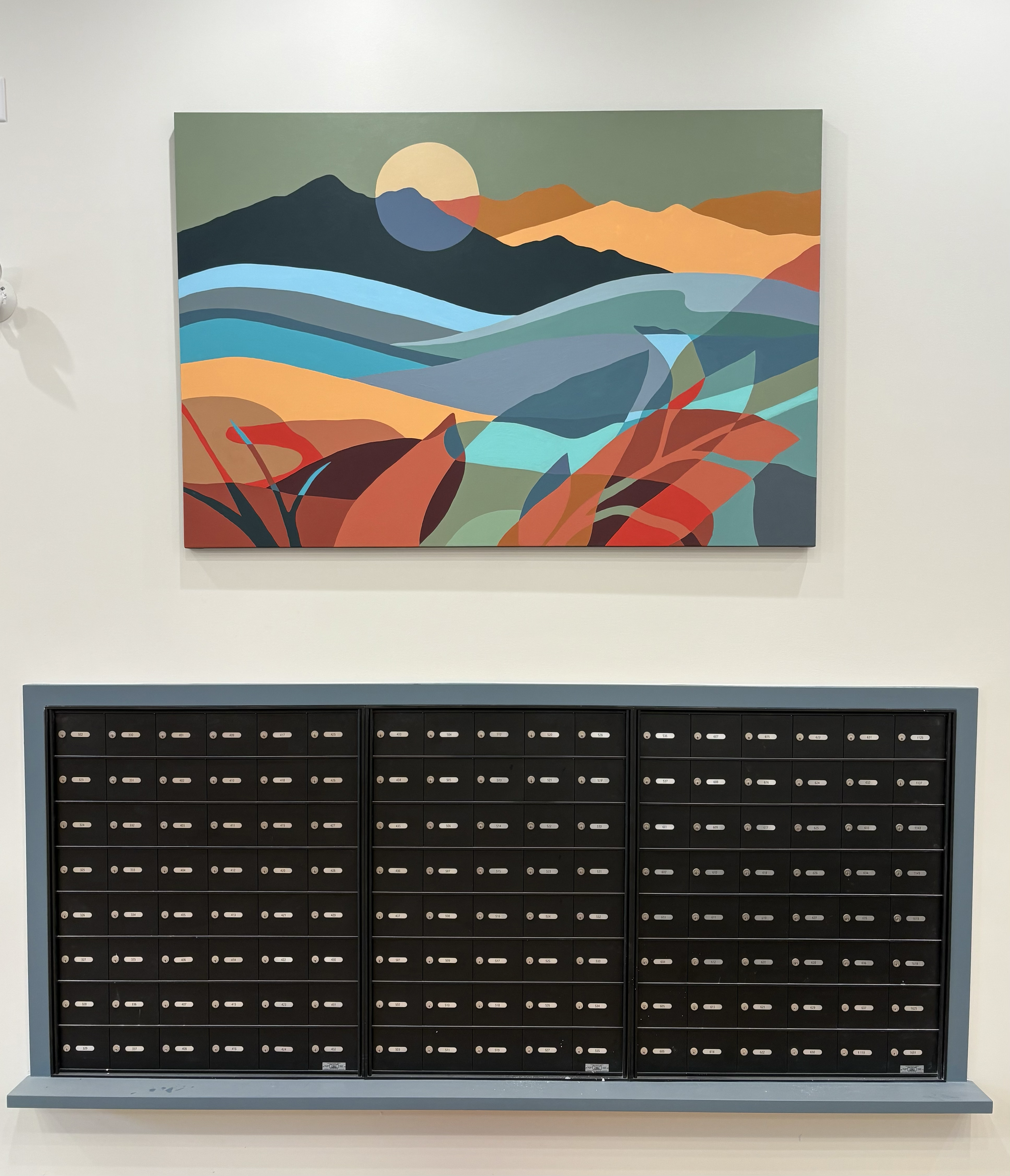 A colorful abstract landscape painting hangs above a set of black mailboxes in a wall.
