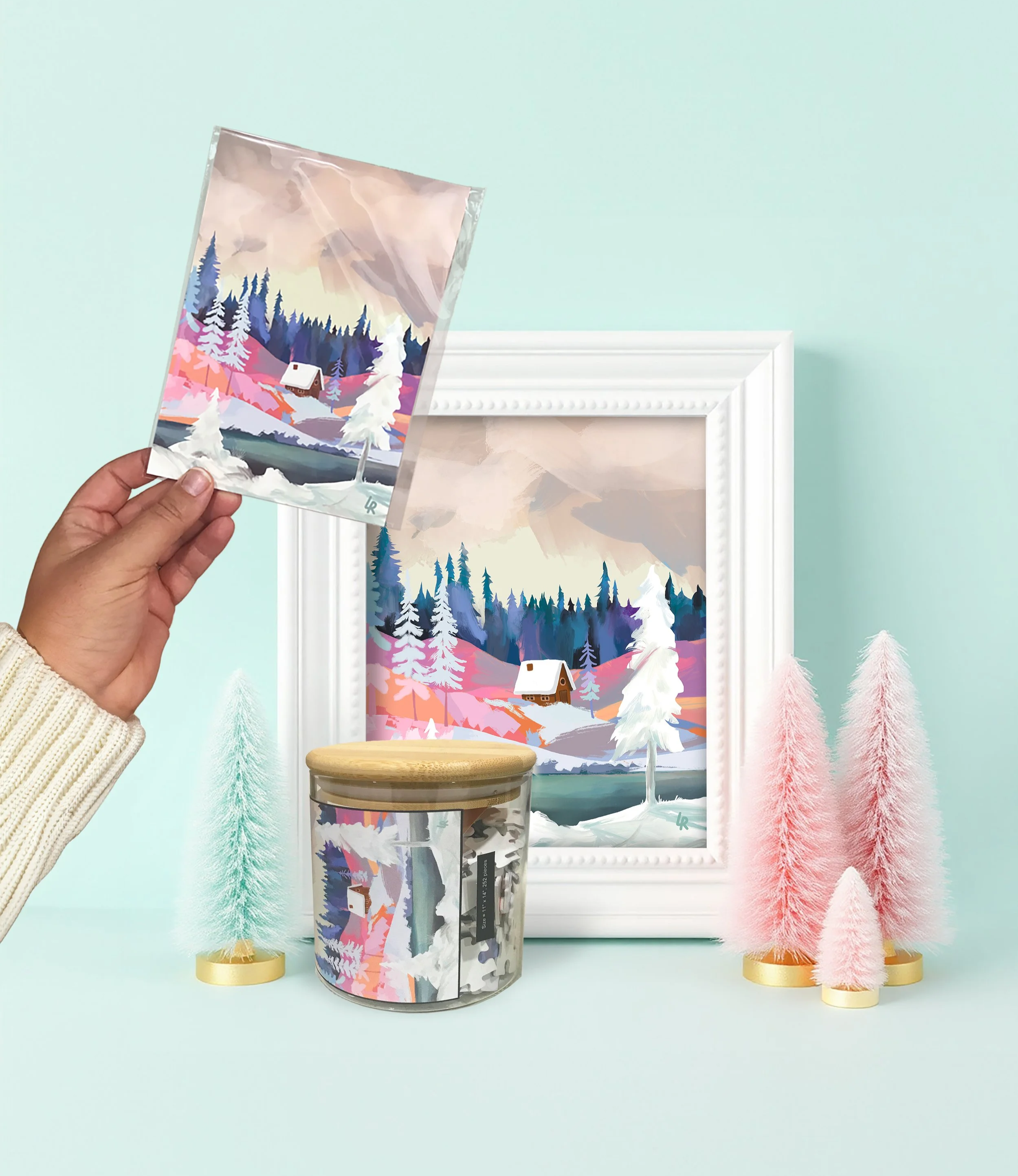 Decorative art scene with a framed winter landscape painting, a candle with a matching winter landscape label, pastel-colored miniature trees, and a person holding a winter landscape card in front of the painting.