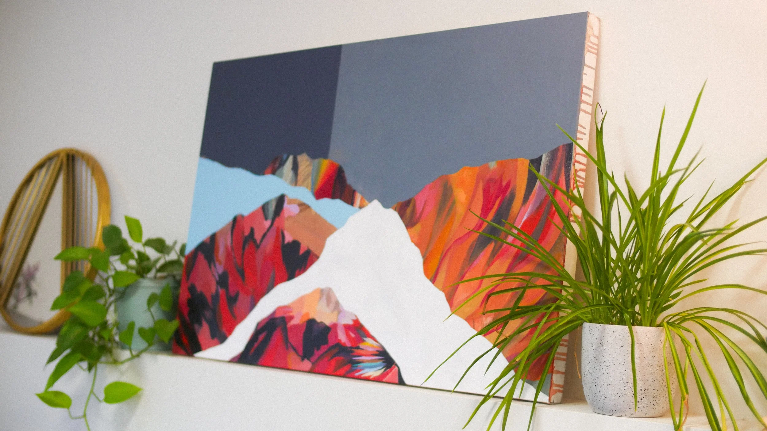 Colorful landscape painting of mountains on a white shelf with potted green plant and decorative mirror in the background.