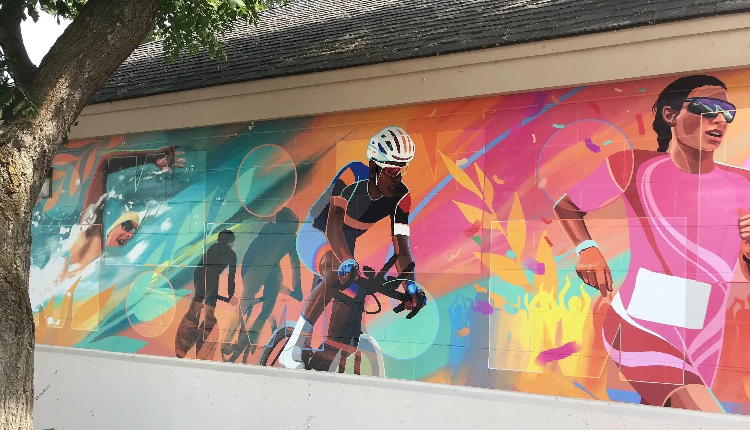 Colorful mural depicting athletes in a race, including a cyclist and runners, with vibrant abstract backgrounds and trees in the foreground.Ironman Canada, Penticton BC, Liz Grace Studio, Liz Dumontet.