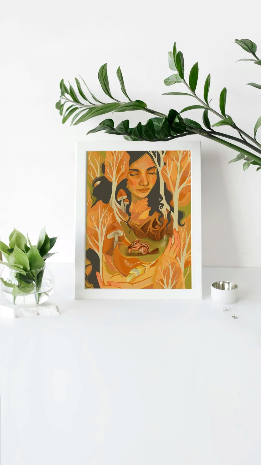 A framed digital artwork of a woman with closed eyes surrounded by orange and brown leaves, with a squirrel and a yellow bird on her hands, placed on a white surface with green plants and a small candle around it.
