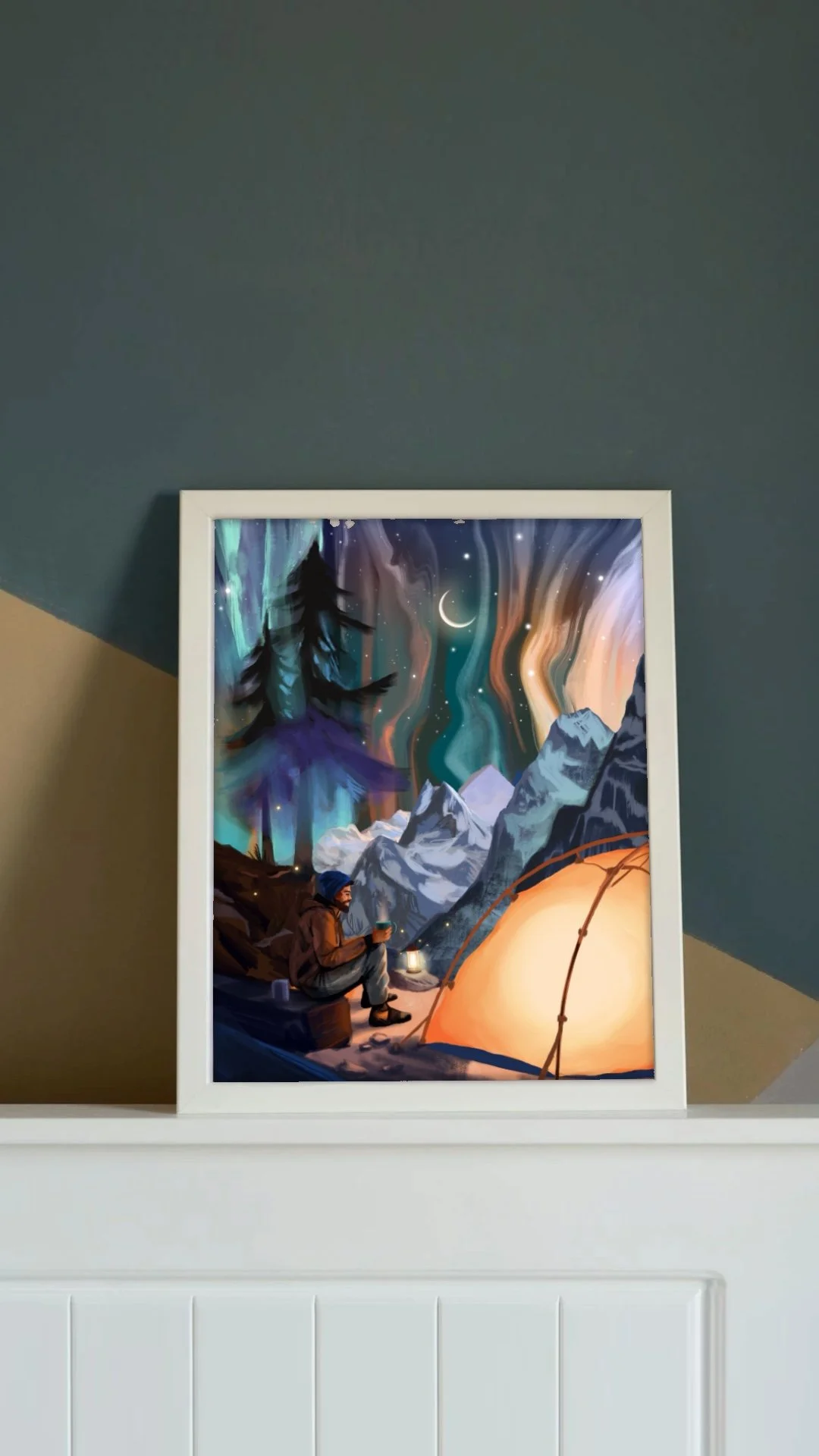 A framed art print of a mountain landscape at night, featuring a colorful aurora, a crescent moon, and a silhouette of a person sitting by a campfire next to a tent.