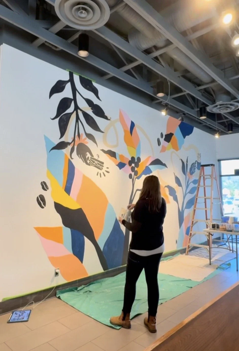 Liz Dumontet, Liz Grace Studio, Artist creating a colorful mural on a white wall in an indoor space, with a ladder and painting supplies nearby. Bright Jenny Coffee, Kelowna British Columbia