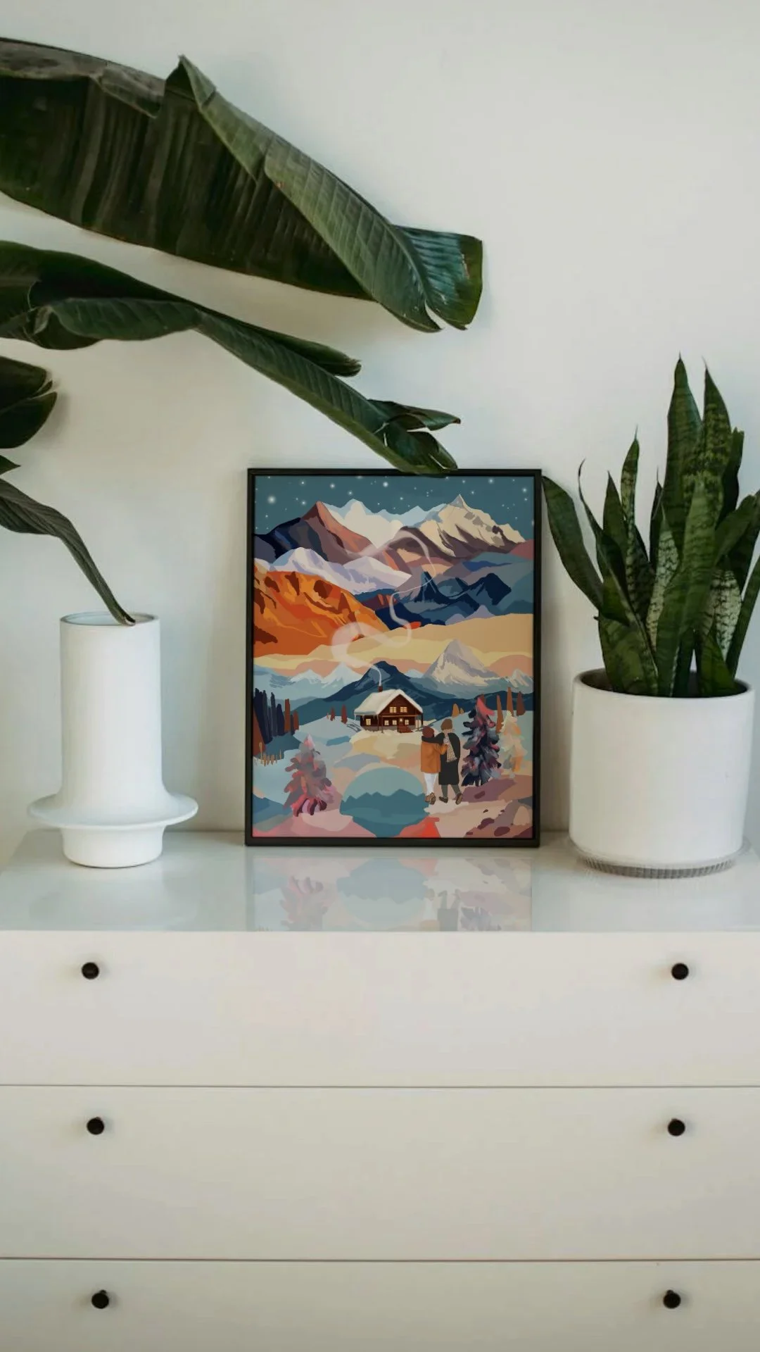 A framed wall art picture featuring a mountain landscape with snow-capped peaks, a sunrise or sunset, a cabin, and two people standing outside, inside a room with tall green plants on either side.