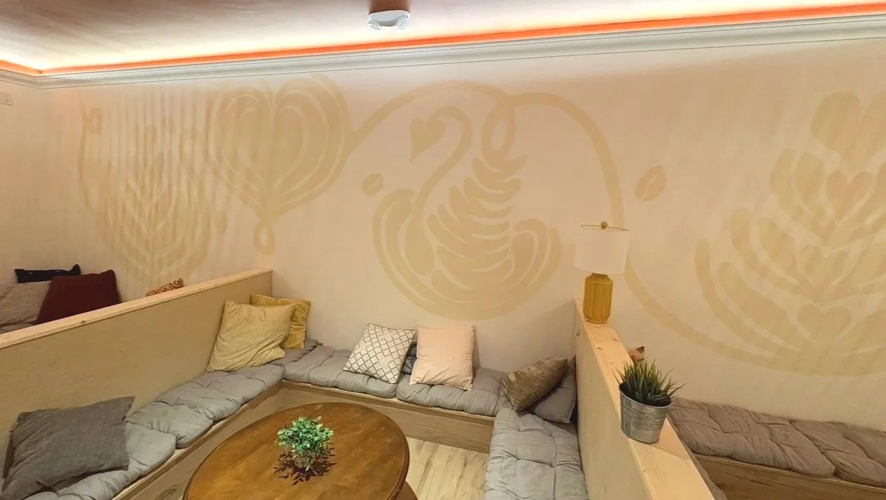 Living room corner with beige wall, decorated with large, stylized, warm-colored floral murals. Sofa seating with various pillows, a small round wooden coffee table, a table lamp, and potted plants. Bright Jenny Coffee, Kelowna BC, Liz Grace Studio,