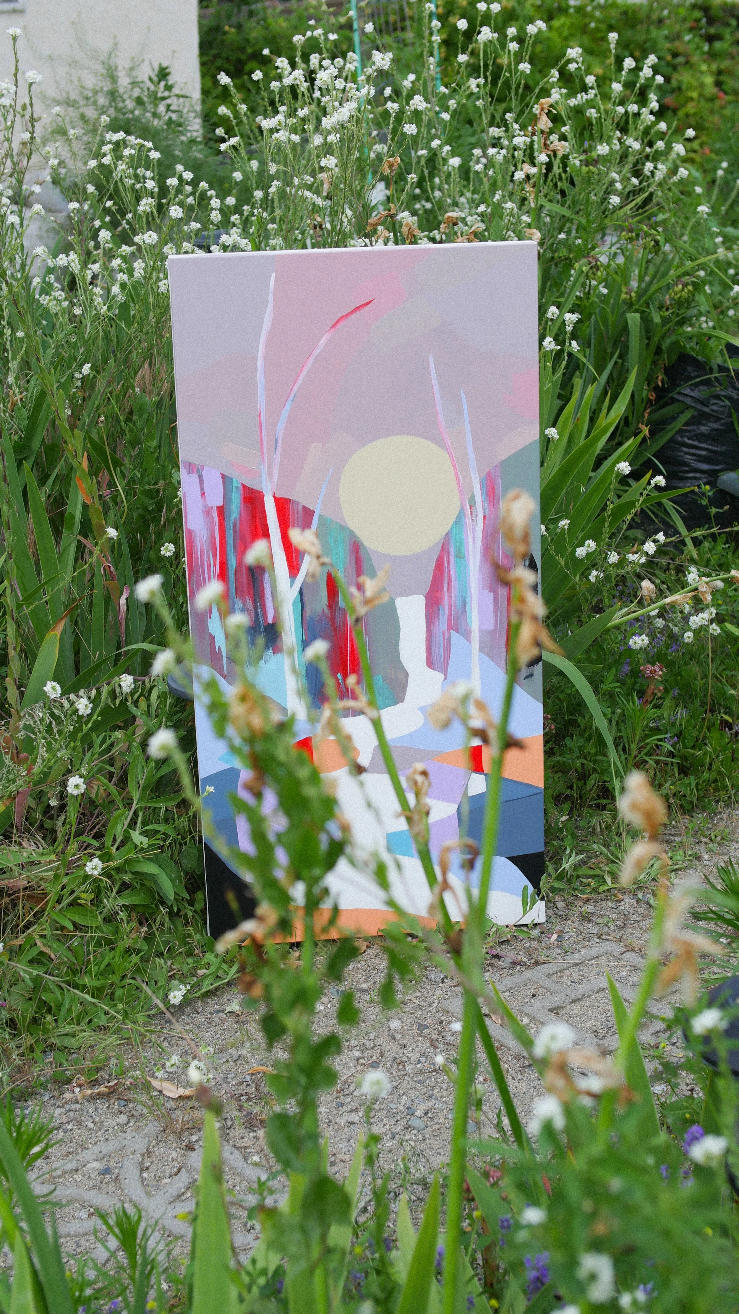 An abstract painting on a canvas, standing outdoors among green plants and small white flowers, featuring pastel colors and shapes.
