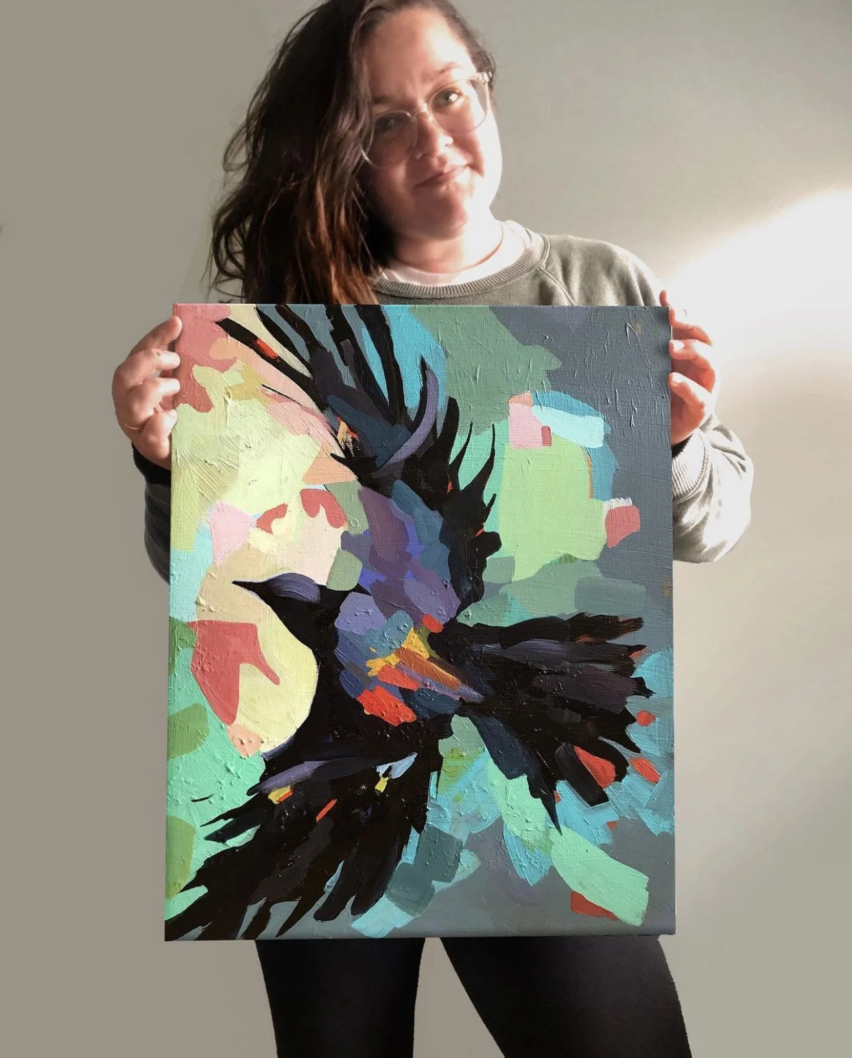Woman with glasses and long hair holding an abstract painting of a bird in flight, with green, blue, purple, red, and black colors.