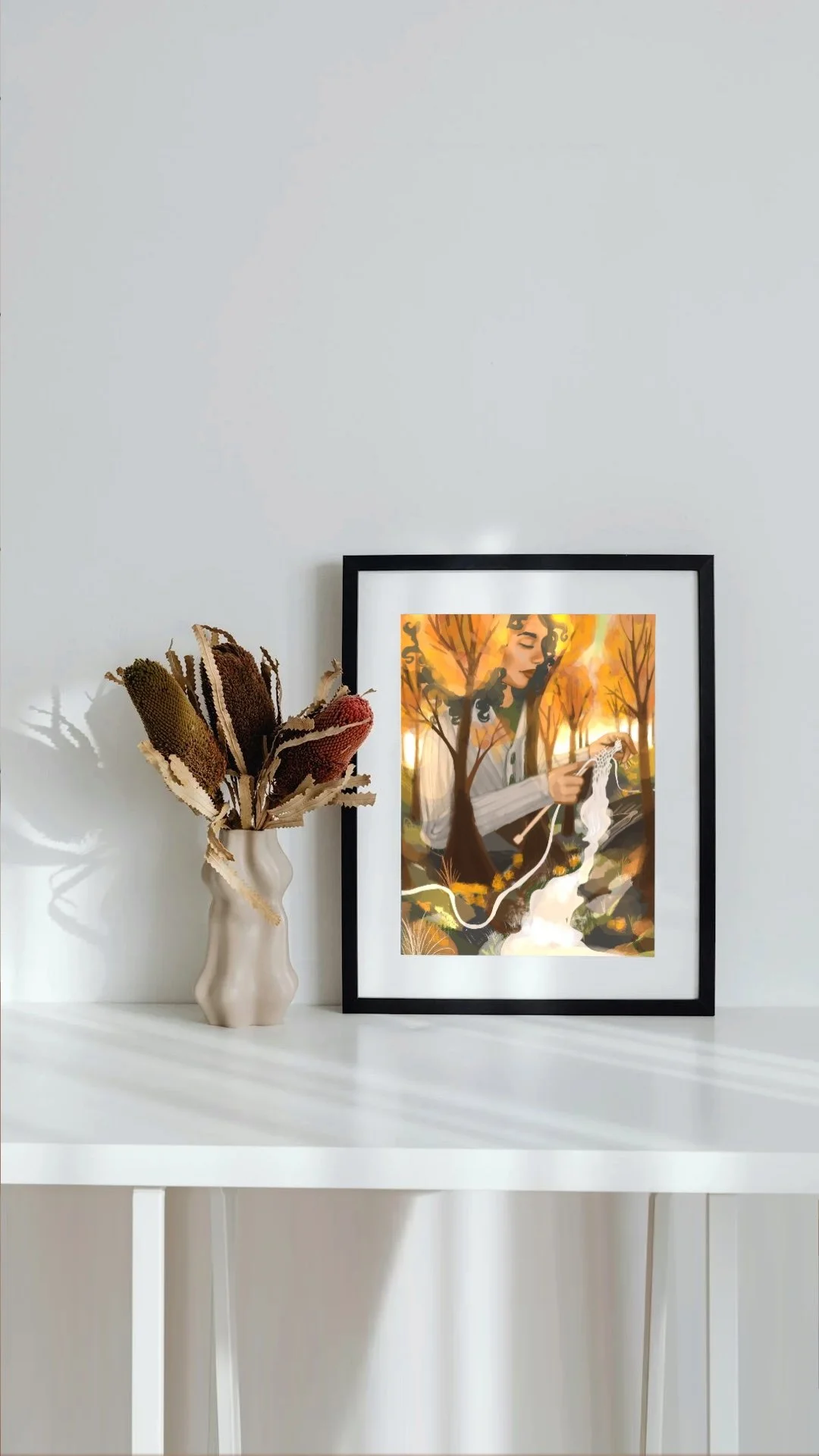 A white table with a small vase holding dried flowers and a framed painting of a woman knitting in an autumn landscape.