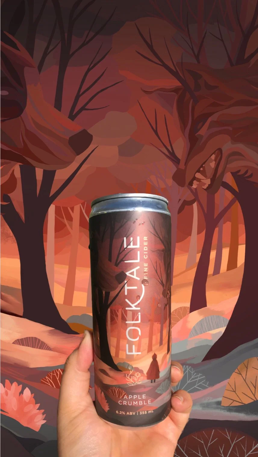 A hand holding a can of Folktale Apple Crumble FINE CIDER in front of a stylized forest background with trees and a wolf.
