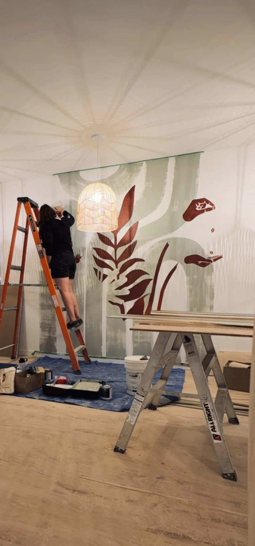 A woman on an orange ladder painting a large botanical mural on a wall, with a hanging light fixture illuminating the scene, and construction tools and wood planks on the floor.
