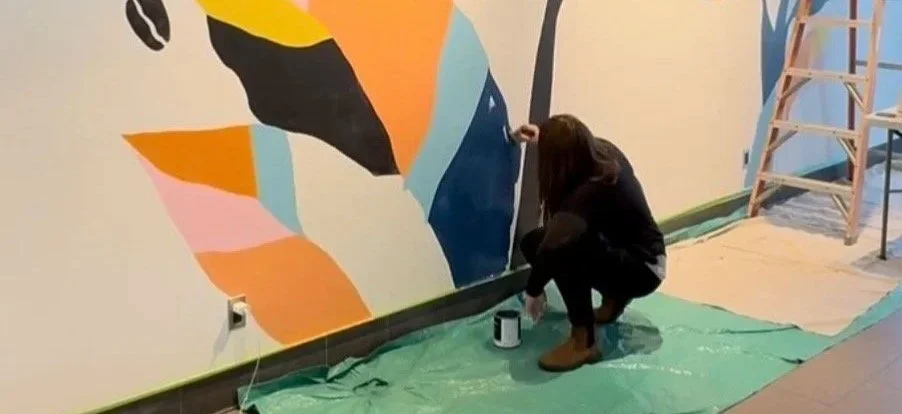A person painting a colorful abstract mural on a wall, kneeling on a green protective sheet with a ladder nearby.