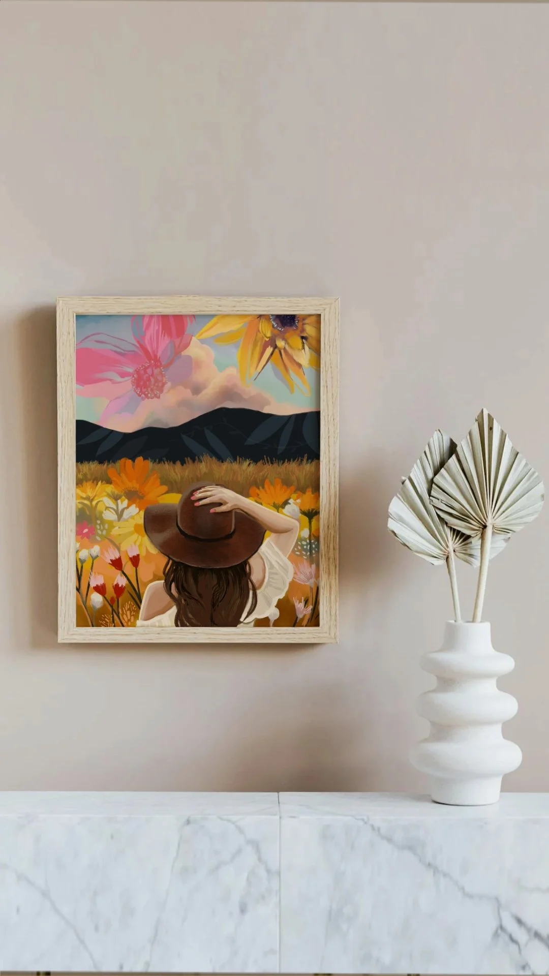 A framed painting of a woman with long brown hair, wearing a large brown hat, standing in a colorful field of flowers with mountains and a partly cloudy sky in the background. A white vase with dried fan leaves is on a marble surface next to the pain