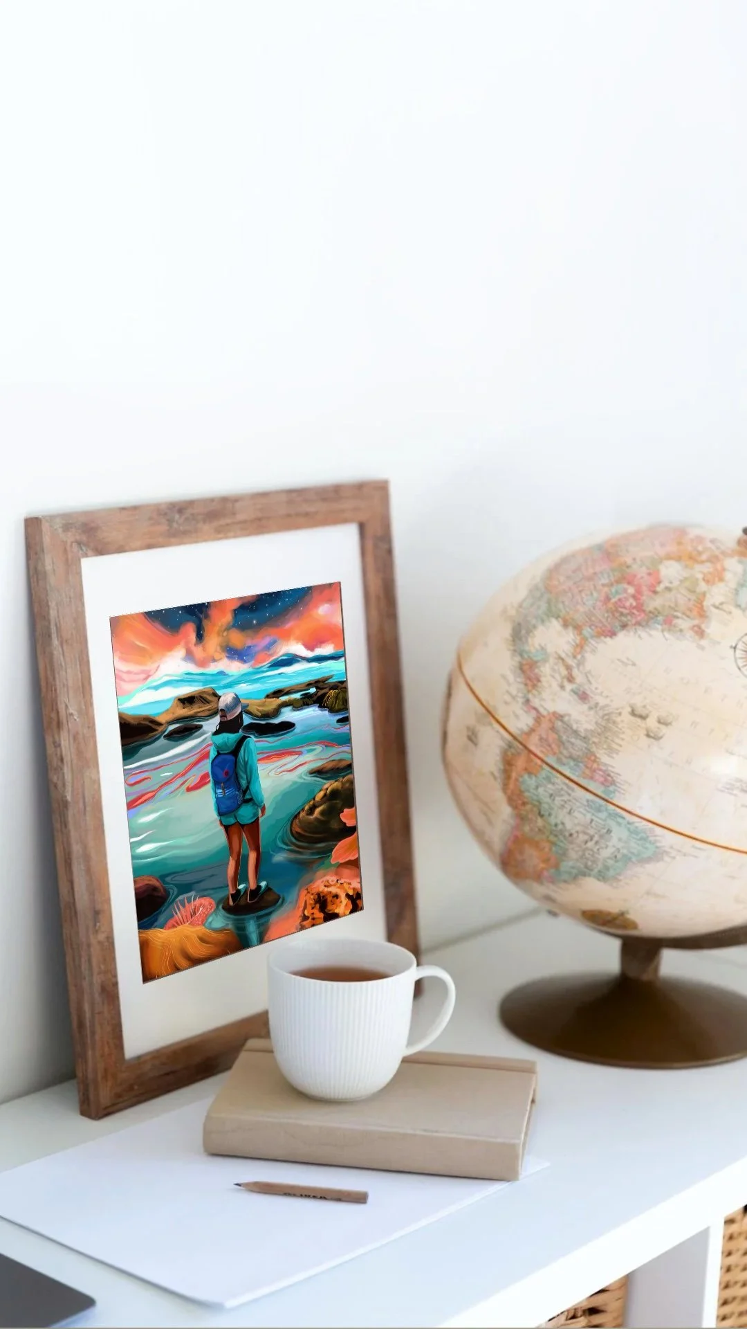 A white desk with a cup of tea, a closed book, and a pencil. On the wall behind, a colorful framed artwork of a person standing near water, with a mountain and sky in the background. To the right, there's a globe.