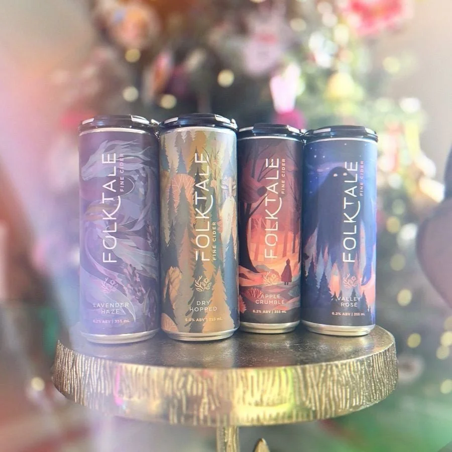 Four cans of Folk Tale cider with colorful, nature-themed labels, placed on a round gold-colored stand in front of a blurred background. Liz Grace Studio, Liz Dumontet.