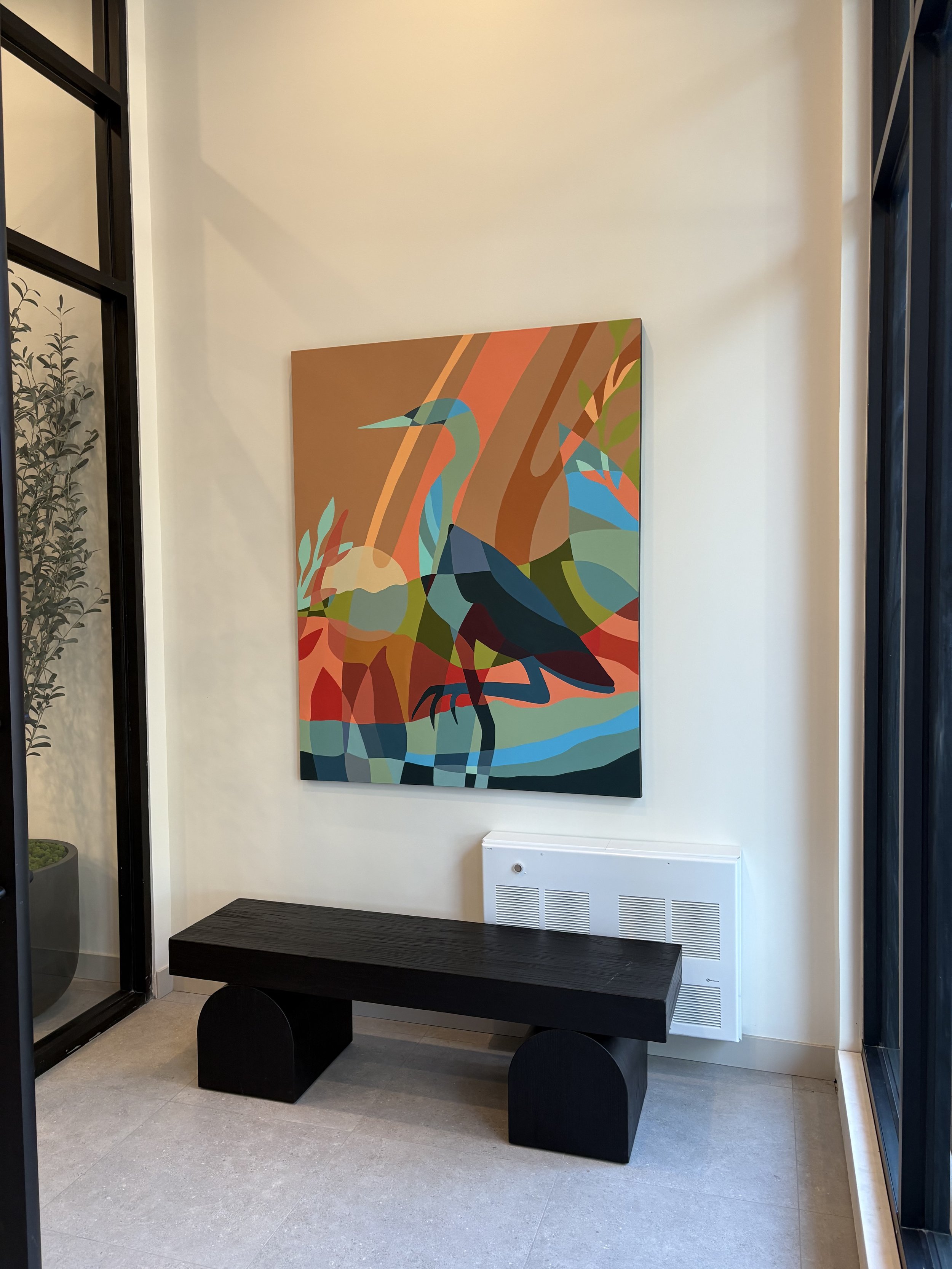 Colorful abstract painting of a heron standing in water, hung on a white wall next to a dark wooden bench and a window.