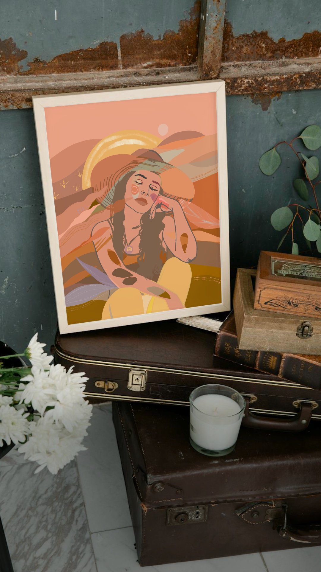 An illustrated art print of a woman with long hair wearing a large sunhat, sitting in a landscape with shades of orange and pink, placed on a vintage brown suitcase, with a candle, a wooden box, and a tree with green leaves nearby.