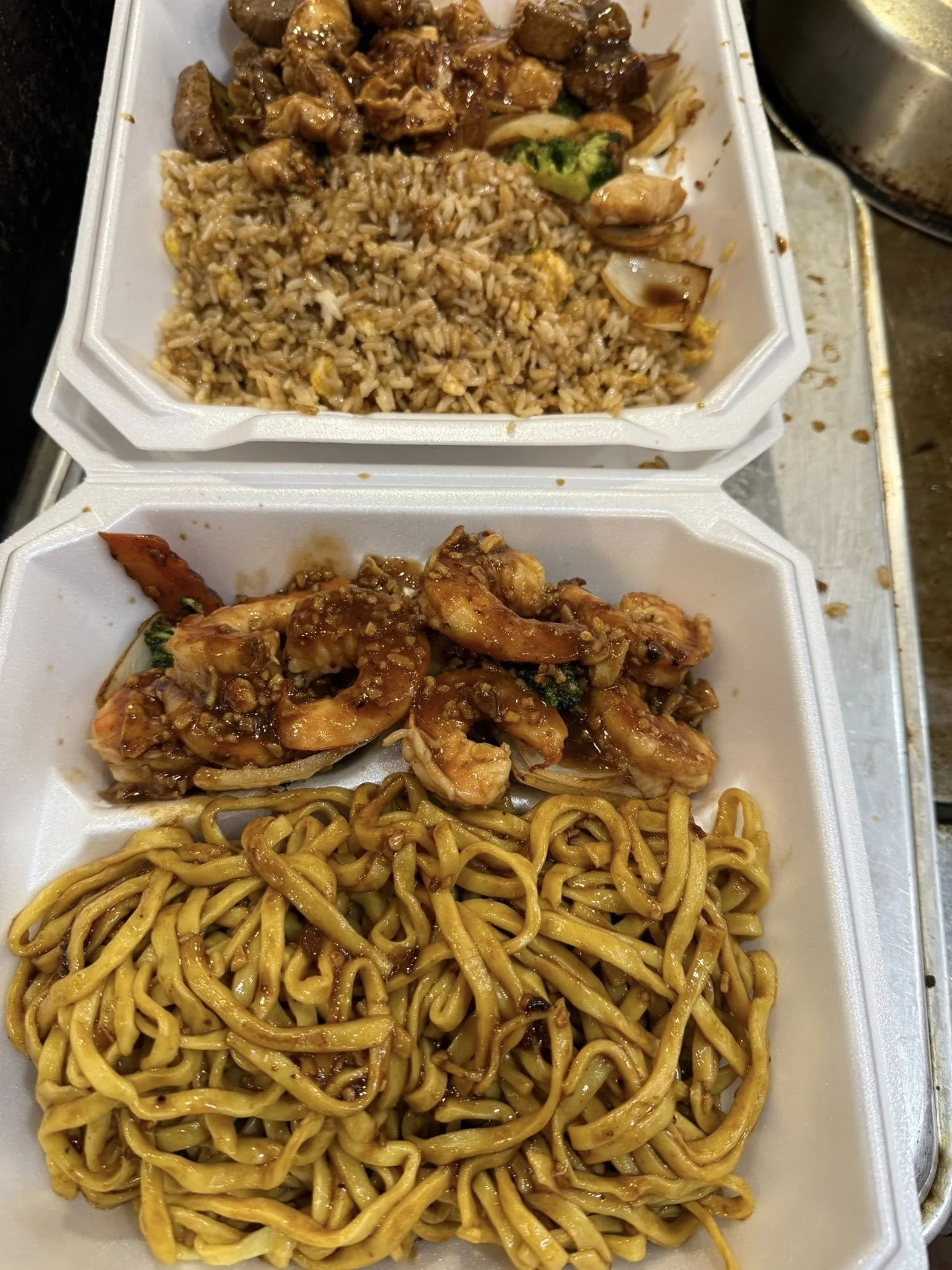 Two takeout styrofoam containers filled with Asian-style food. The top container has fried rice with egg, chunks of beef, broccoli, and onions. The bottom container features stir-fried noodles topped with shrimp, vegetables, and a dark sauce.