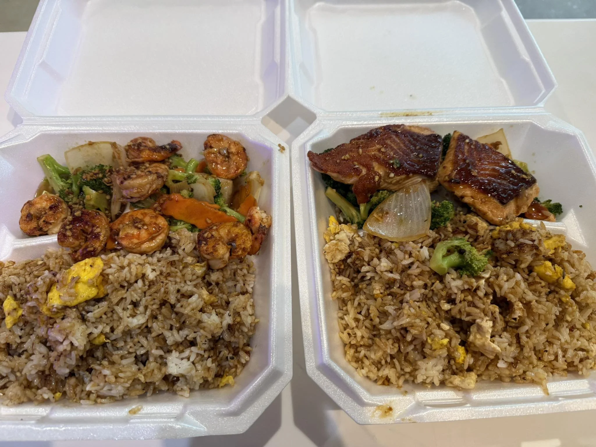 Two foam takeout containers filled with fried rice, vegetables, and grilled chicken or shellfish.