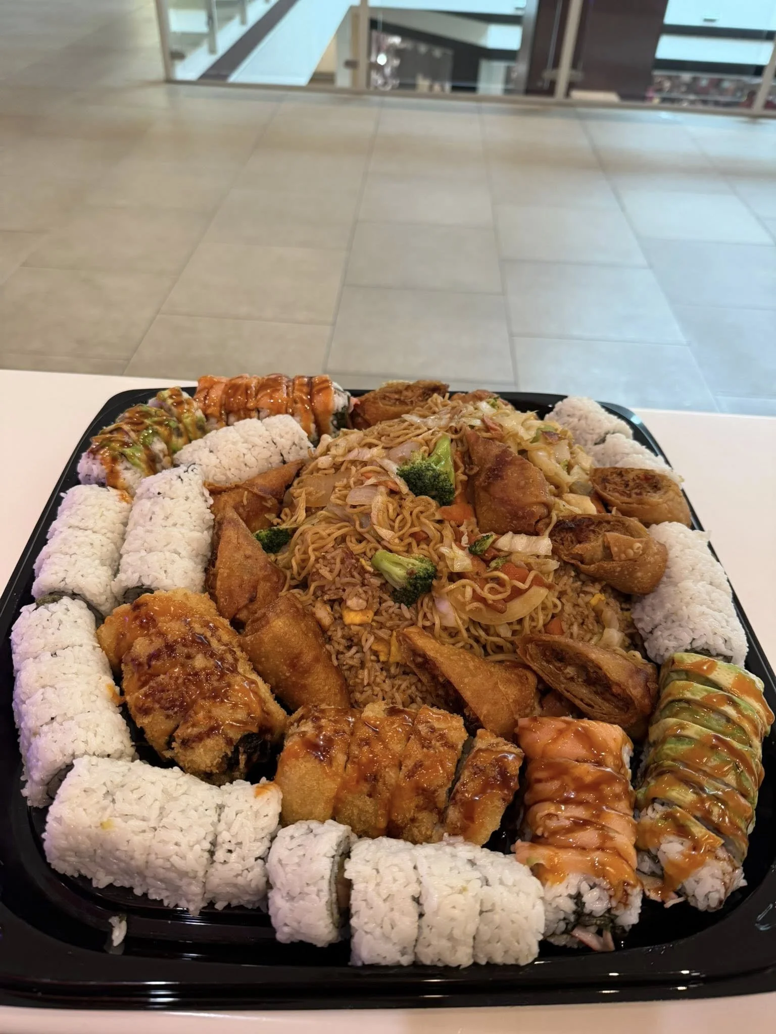A black tray filled with assorted sushi rolls and Chinese food like fried rice, spring rolls, and noodles.