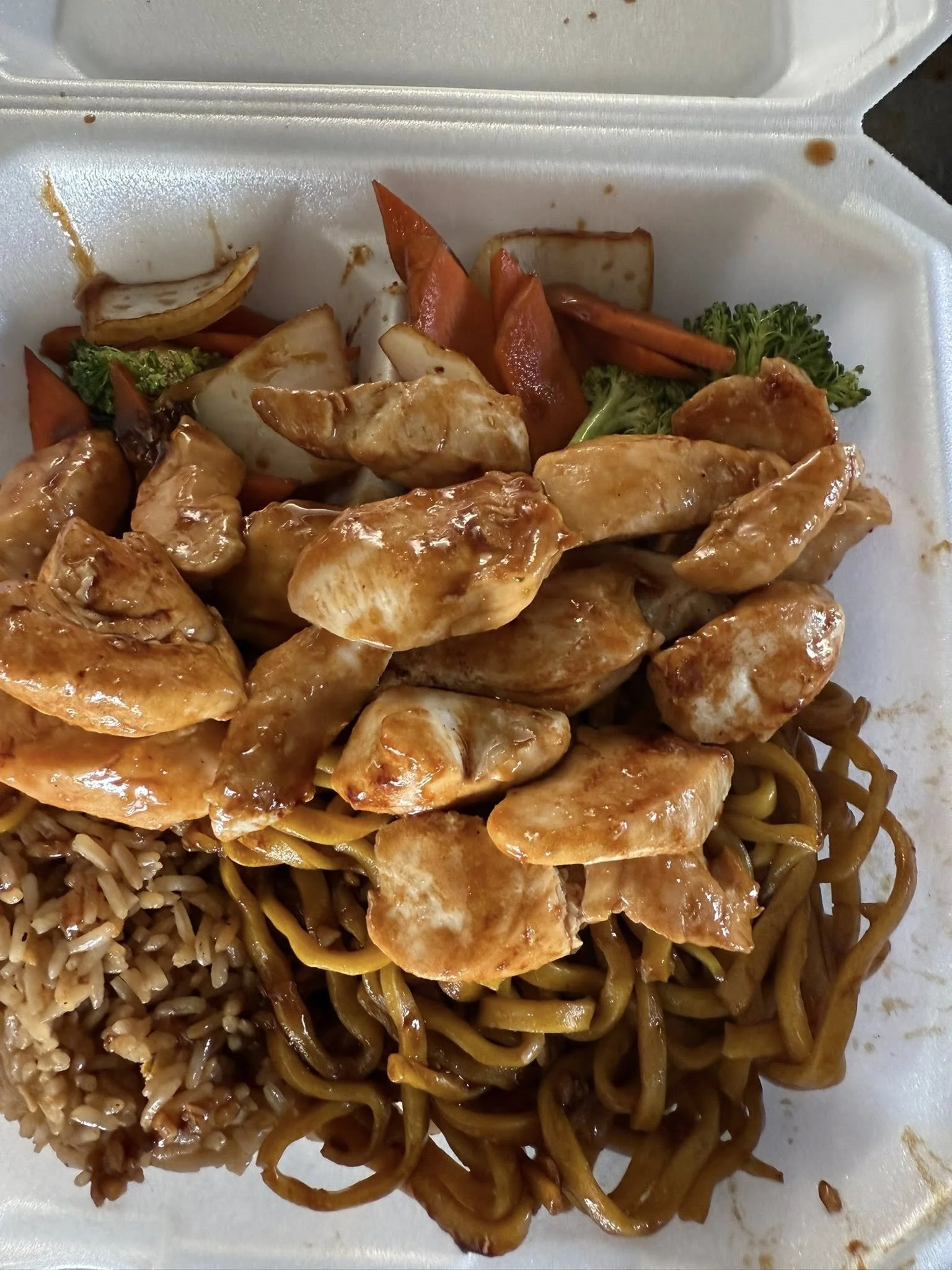 Takeout container with Asian stir-fried chicken, noodles, rice, and mixed vegetables including broccoli, carrots, and onions.