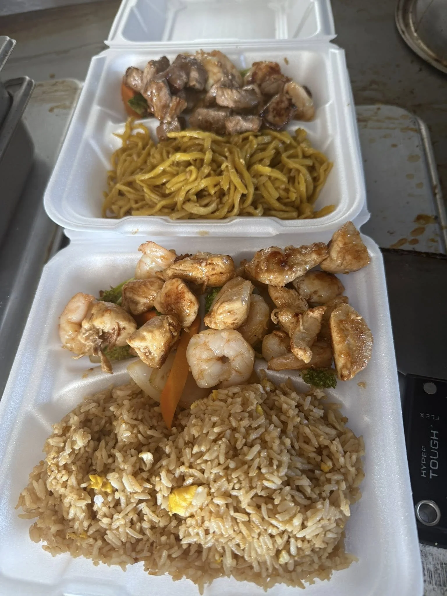 Two styrofoam takeout containers with Asian-style food. The top container has noodles and mixed vegetables with chicken, the bottom container has fried rice with vegetables, shrimp, and pieces of chicken.