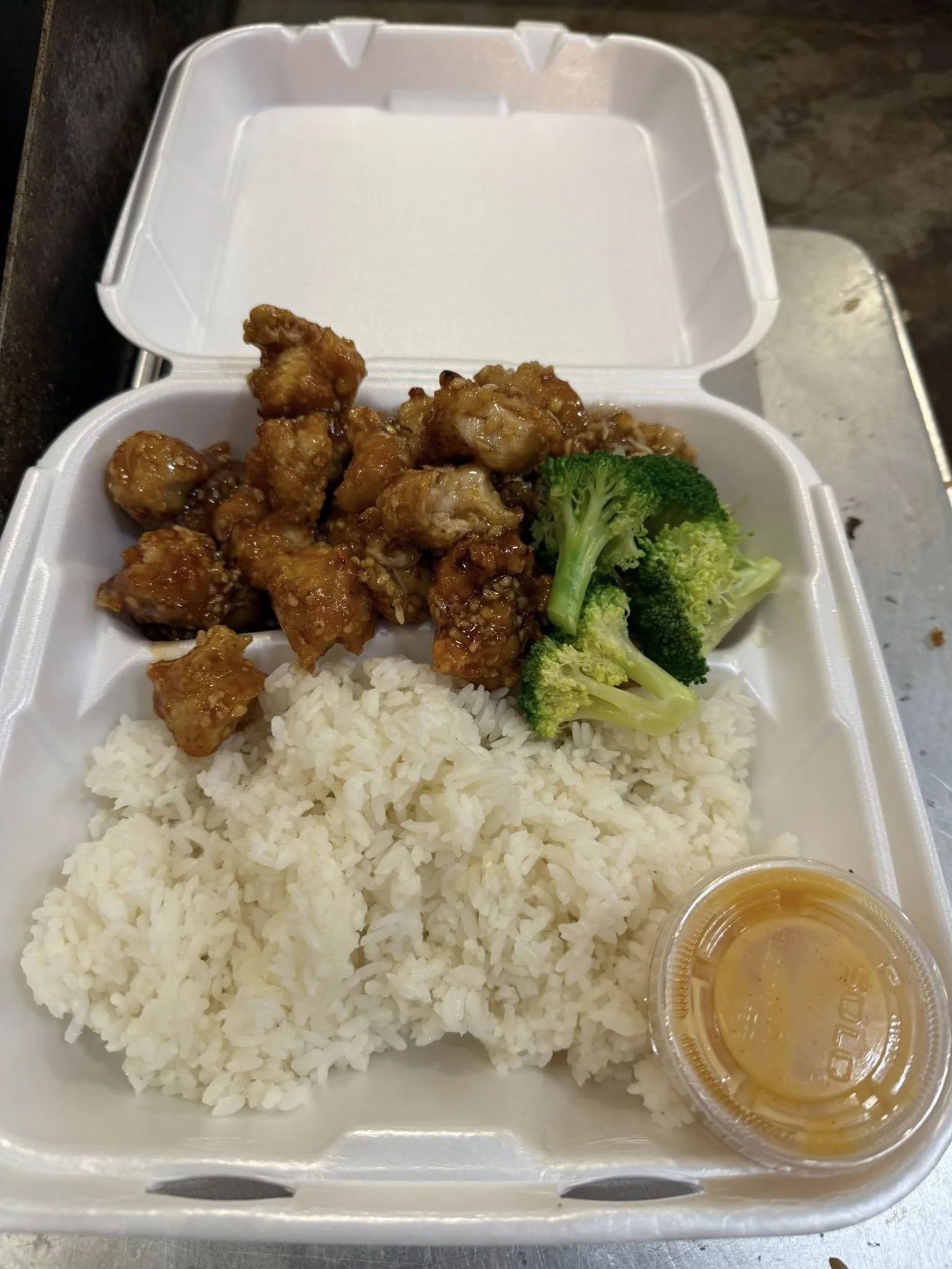 Takeout container with fried chicken pieces, steamed white rice, broccoli florets, and a small cup of sauce.