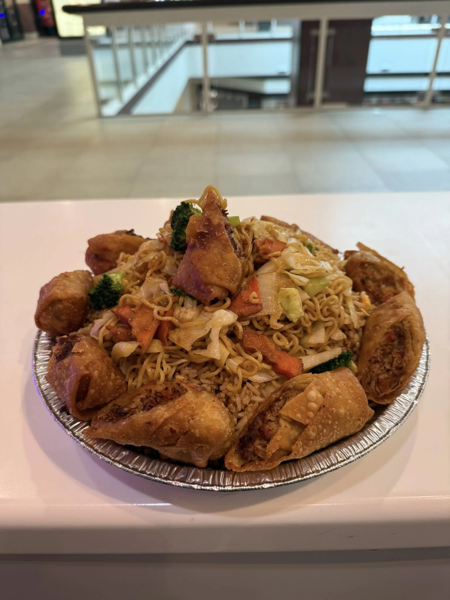 Fried spring rolls surrounding a bowl of fried rice with vegetables and chicken in a takeout container.