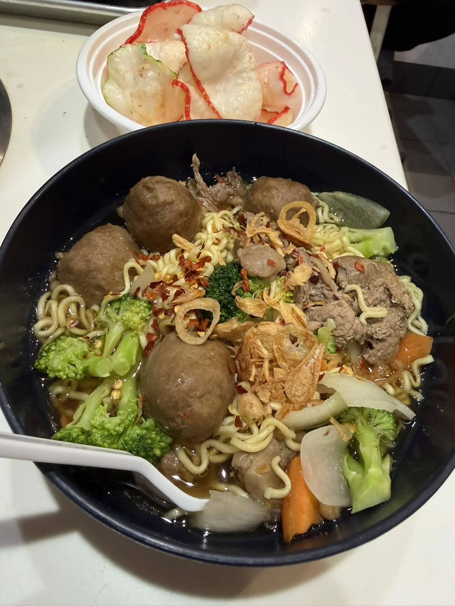 Bowl of Asian-style noodle soup with meatballs, beef slices, vegetables, and fried onions, alongside a small dish of potato chips with red and green peppers.