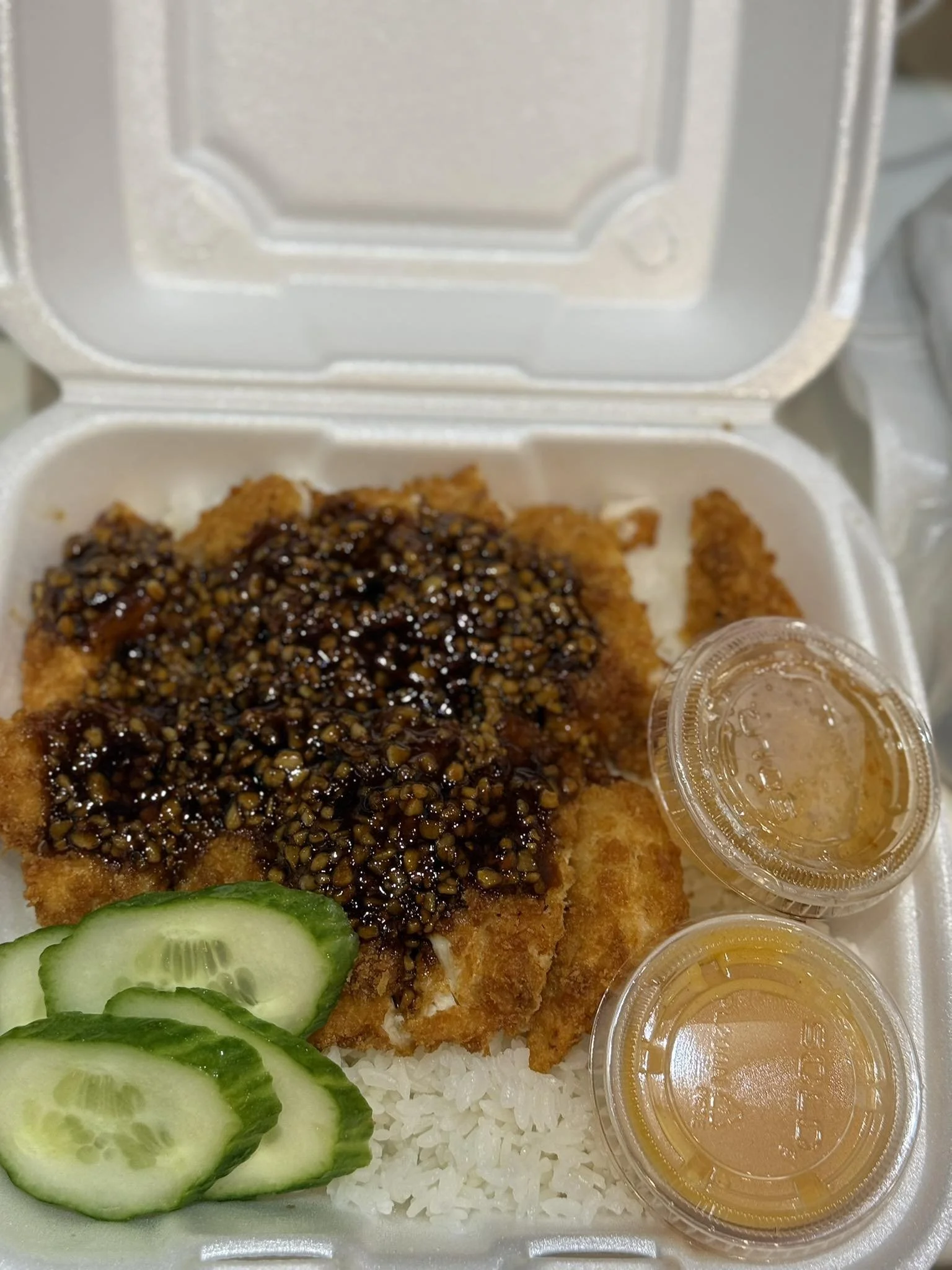 Fried fish fillet with black bean sauce, white rice, sliced cucumbers, and two small containers of sauce in a takeout box.