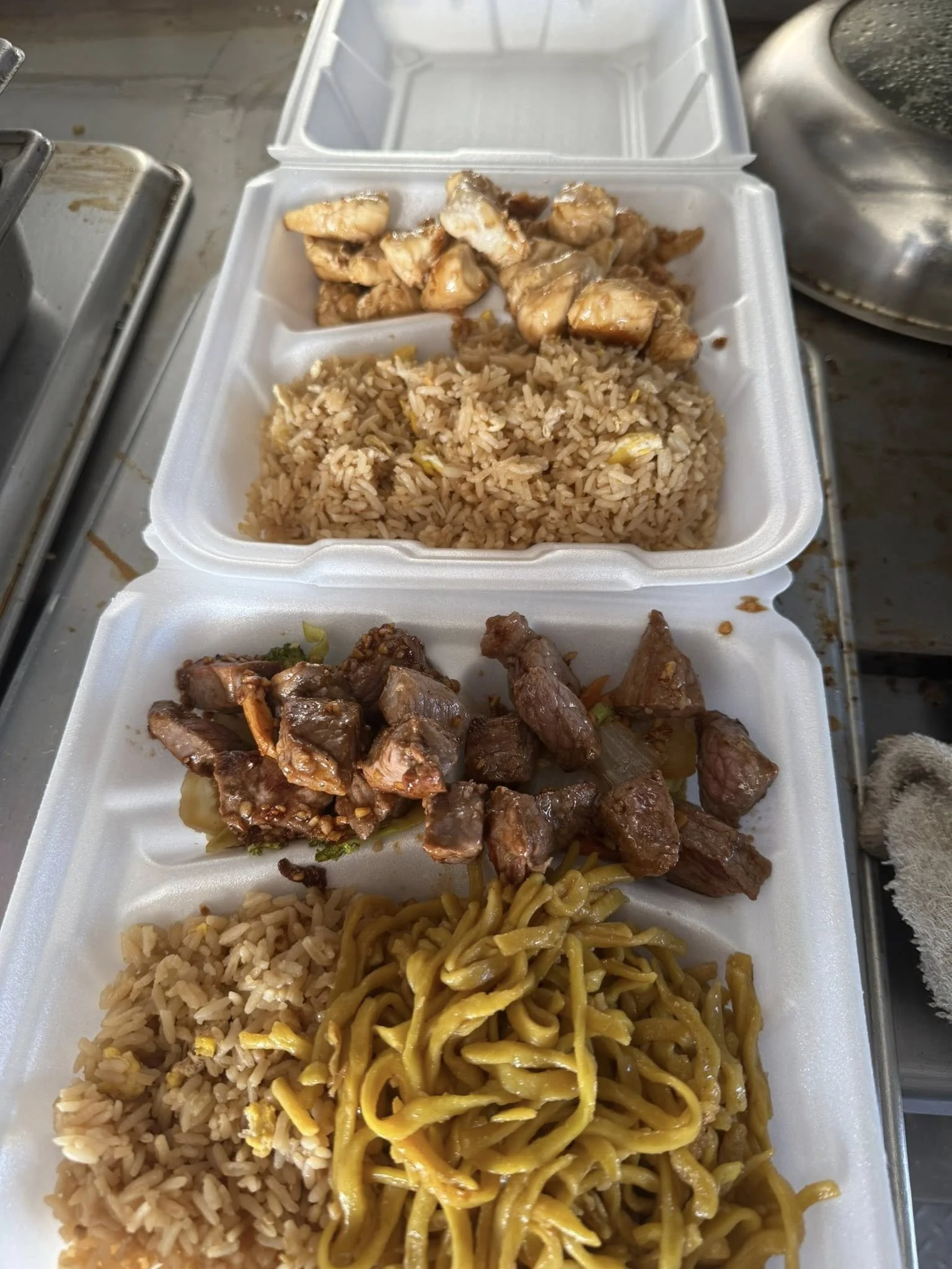 A styrofoam takeout container with three compartments filled with Asian-style food, including rice, stir-fried noodles, chicken, and beef, on a metal surface.