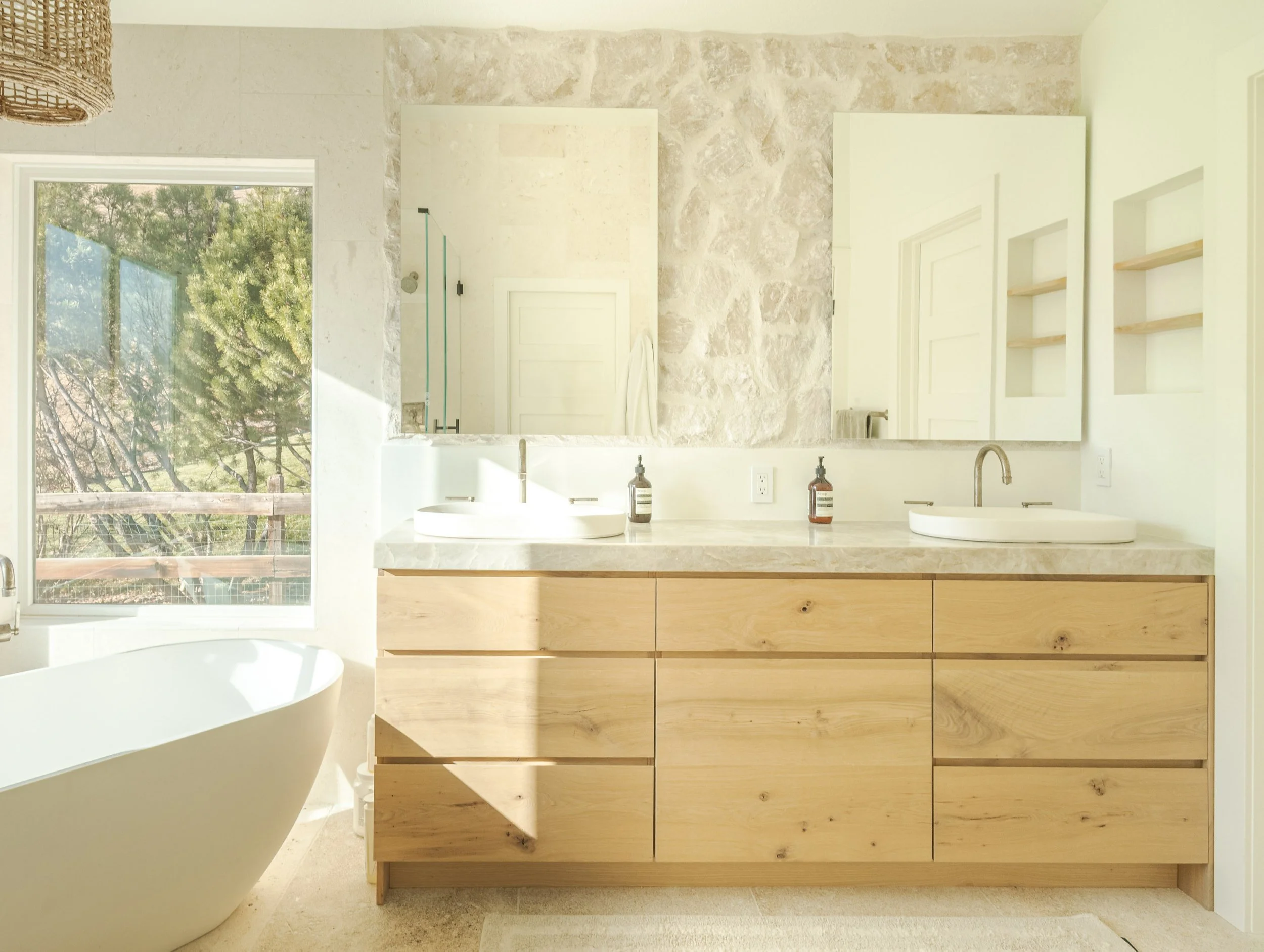 Bright bathroom featuring a large mirror above a wood vanity with two white vessel sinks, a freestanding bathtub by a window with a view of a tree, and a stone wall with a door and shelves reflected in the mirror.