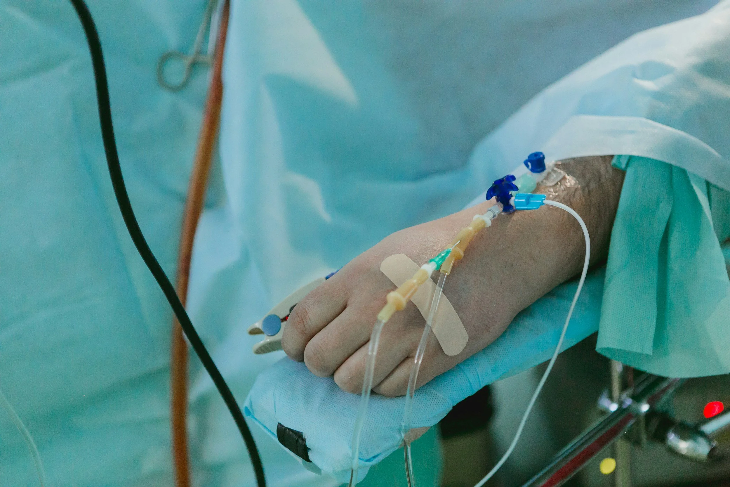 Close-up of a patient's hand with IV lines and monitors attached, lying on a hospital bed in an operating room.