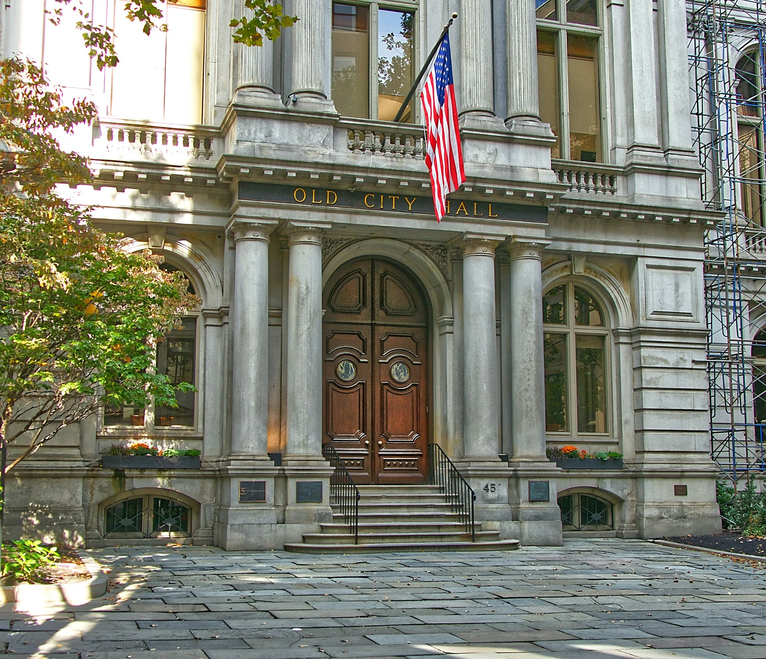 Front entrance of the Old City Hall building with a large wooden door, stone steps, columns, a balcony with an American flag, and the building's name above the doorway, numbered 45.