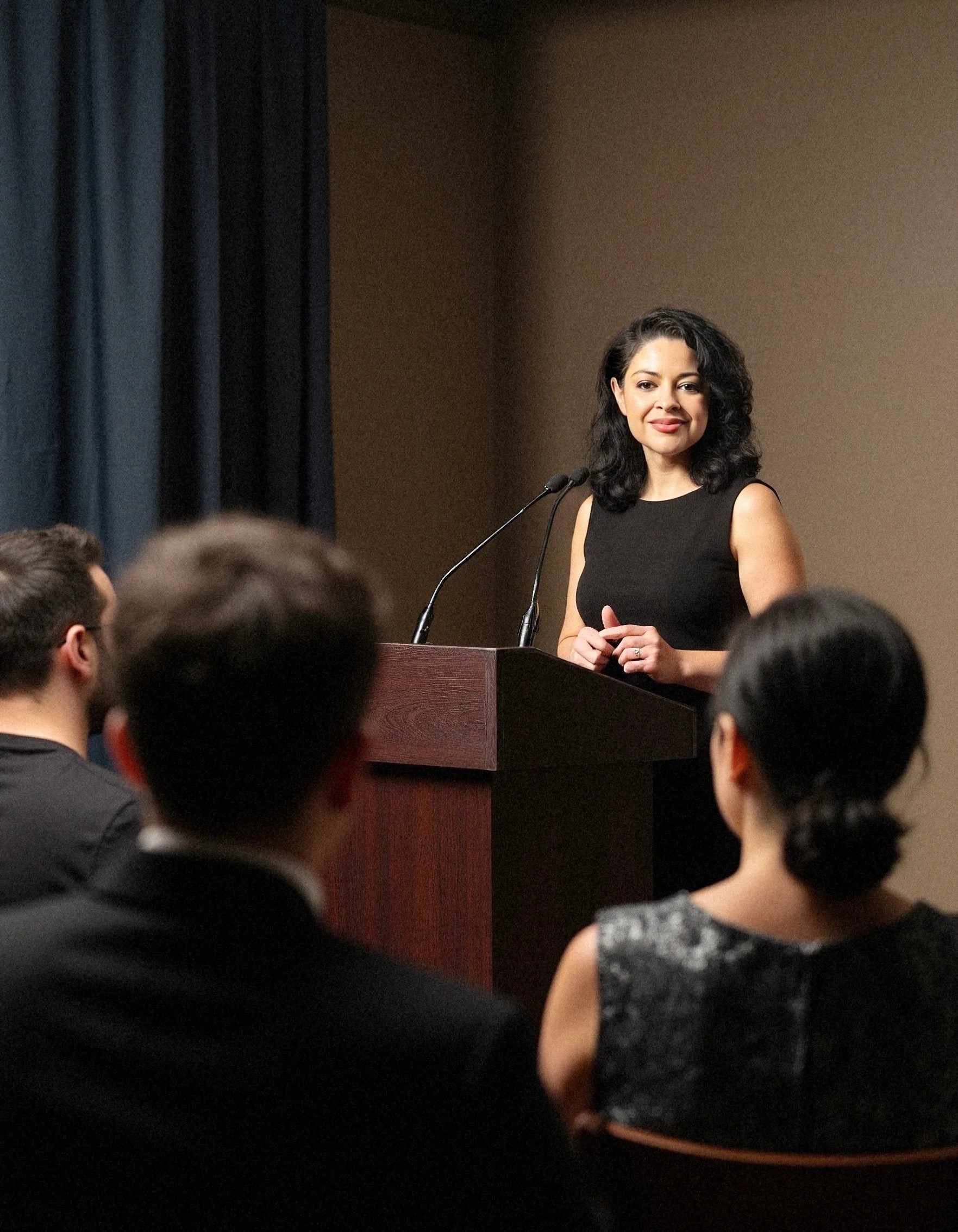 A woman with dark, curly hair and wearing a black sleeveless dress stands at a podium, speaking into two microphones during a formal event, with audience members visible in the foreground.