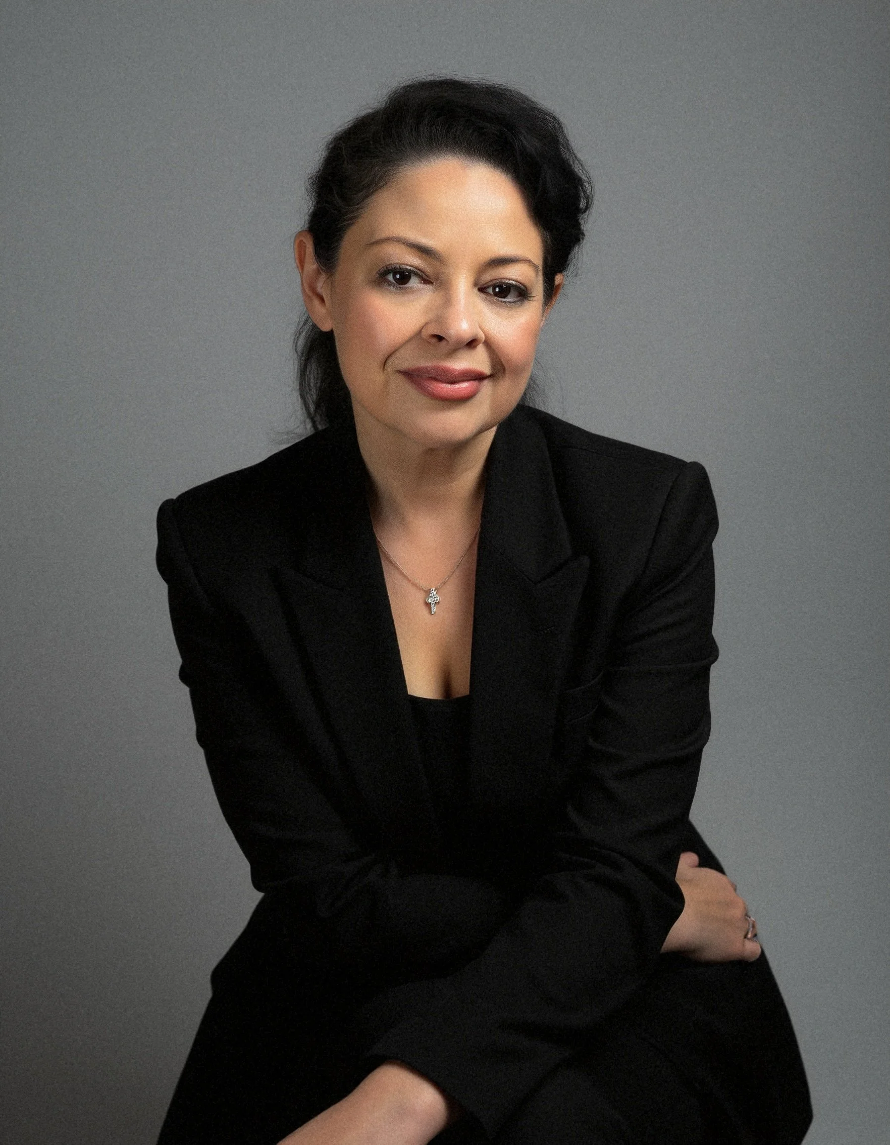 A woman with dark hair and light skin wearing a black blazer and a necklace with a cross pendant sitting against a gray background.