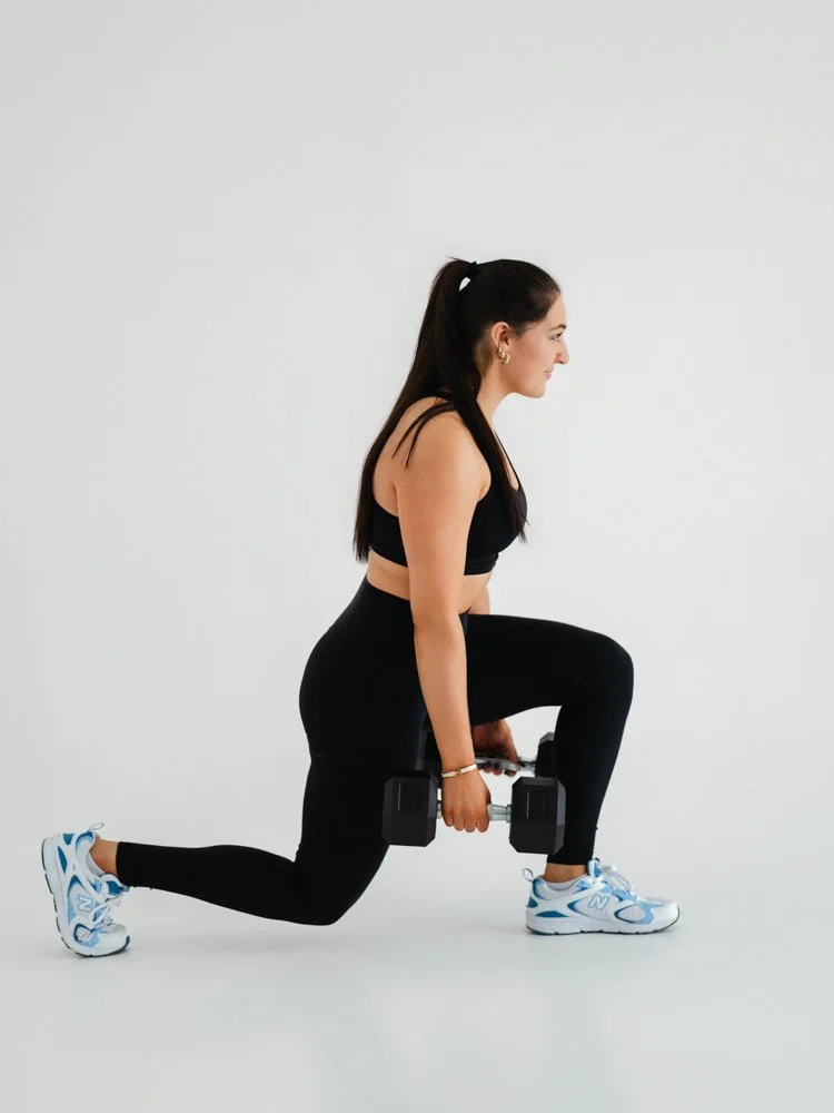 A woman in black workout clothes and sneakers performs a lunge exercise holding dumbbells against a plain white background.